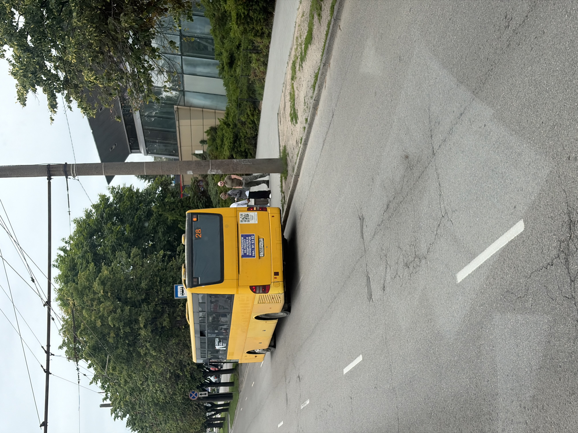 A yellow bus is stopped at a bus stop on a street in Kaliningrad, Russia. The bus has the number 28 displayed on its rear. Three people are standing near the bus, one appears to be a woman looking at her phone, and two men are standing and conversing. The bus stop has a blue sign with a white pictogram of a bus. In the background, there is a modern building with large windows and a sloped roof, surrounded by lush green trees and bushes. Overhead, there are numerous power lines crisscrossing the sky. The street is asphalt with painted white lines marking the lanes. The day appears overcast.