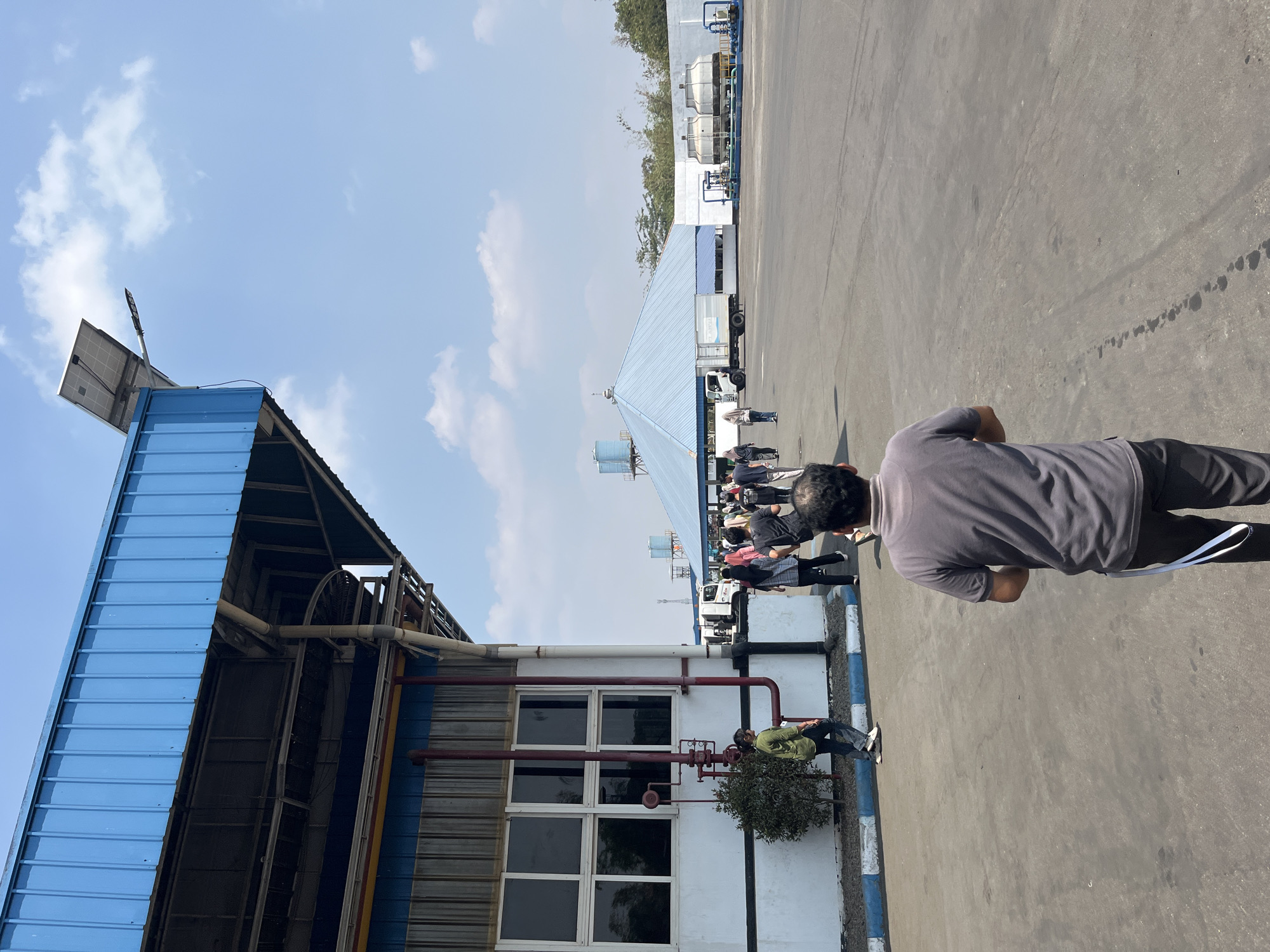 In the city of Pelang, Indonesia, a man is walking away from the camera on a paved industrial area. He is wearing a grey polo shirt and dark pants. Several other people are walking in the distance towards a large, light blue building with a peaked roof. A white truck is parked near the building, and there are industrial structures visible in the background, including cooling towers. The sky is blue with some white clouds.