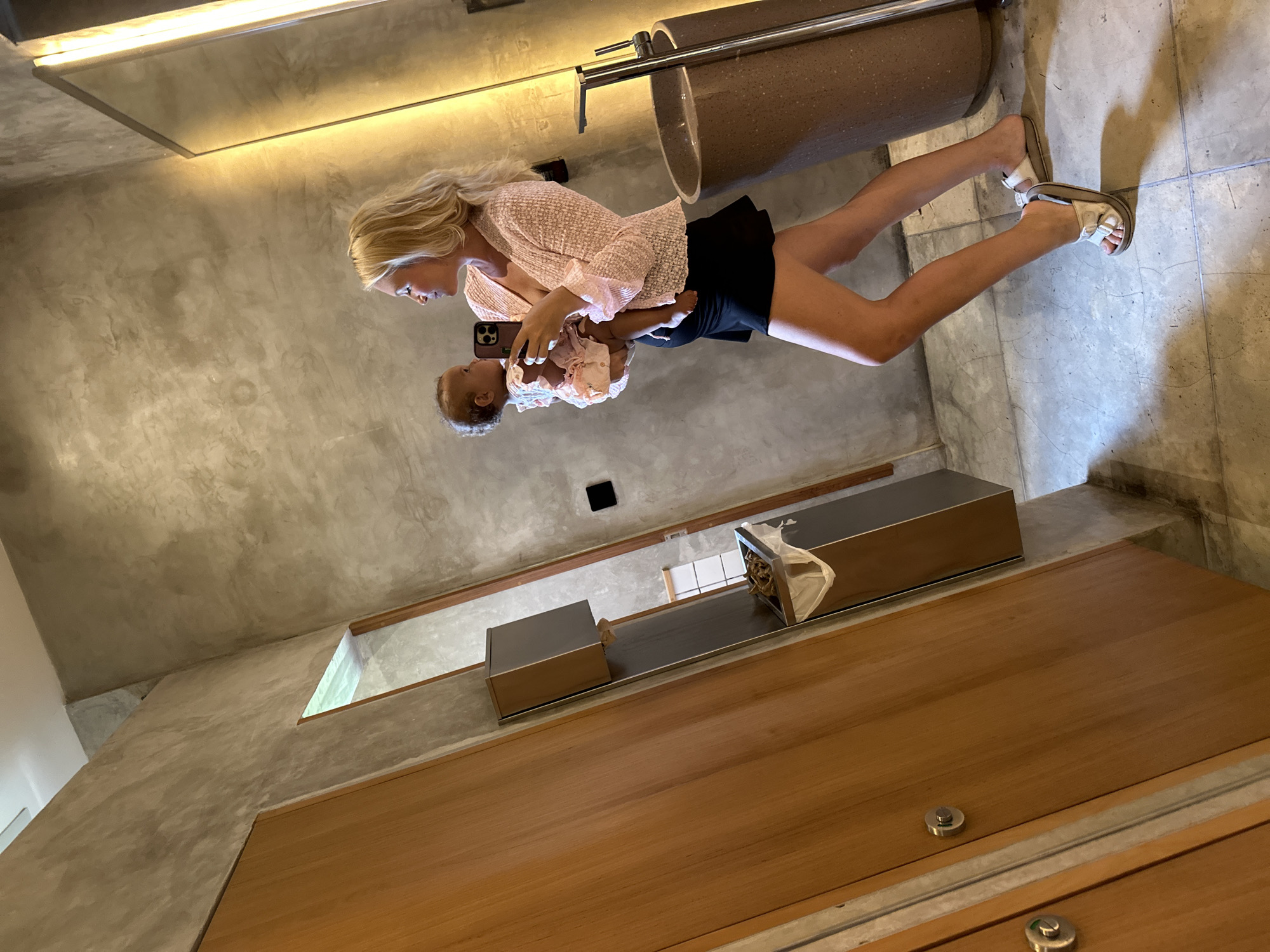In a modern bathroom with textured concrete walls and wooden doors, a blonde woman is taking a mirror selfie while holding a baby. The woman is wearing a pink, textured, open-front cardigan over a black skirt. She is looking at her reflection and at the baby she holds in her left arm. The baby is wearing a light-colored, ruffled dress and is looking towards the woman. To the right of the woman, a large, illuminated mirror hangs on the wall above a textured brown sink with a chrome faucet. The floor is tiled with grey concrete. The scene appears to be in North Kuta, Indonesia.
