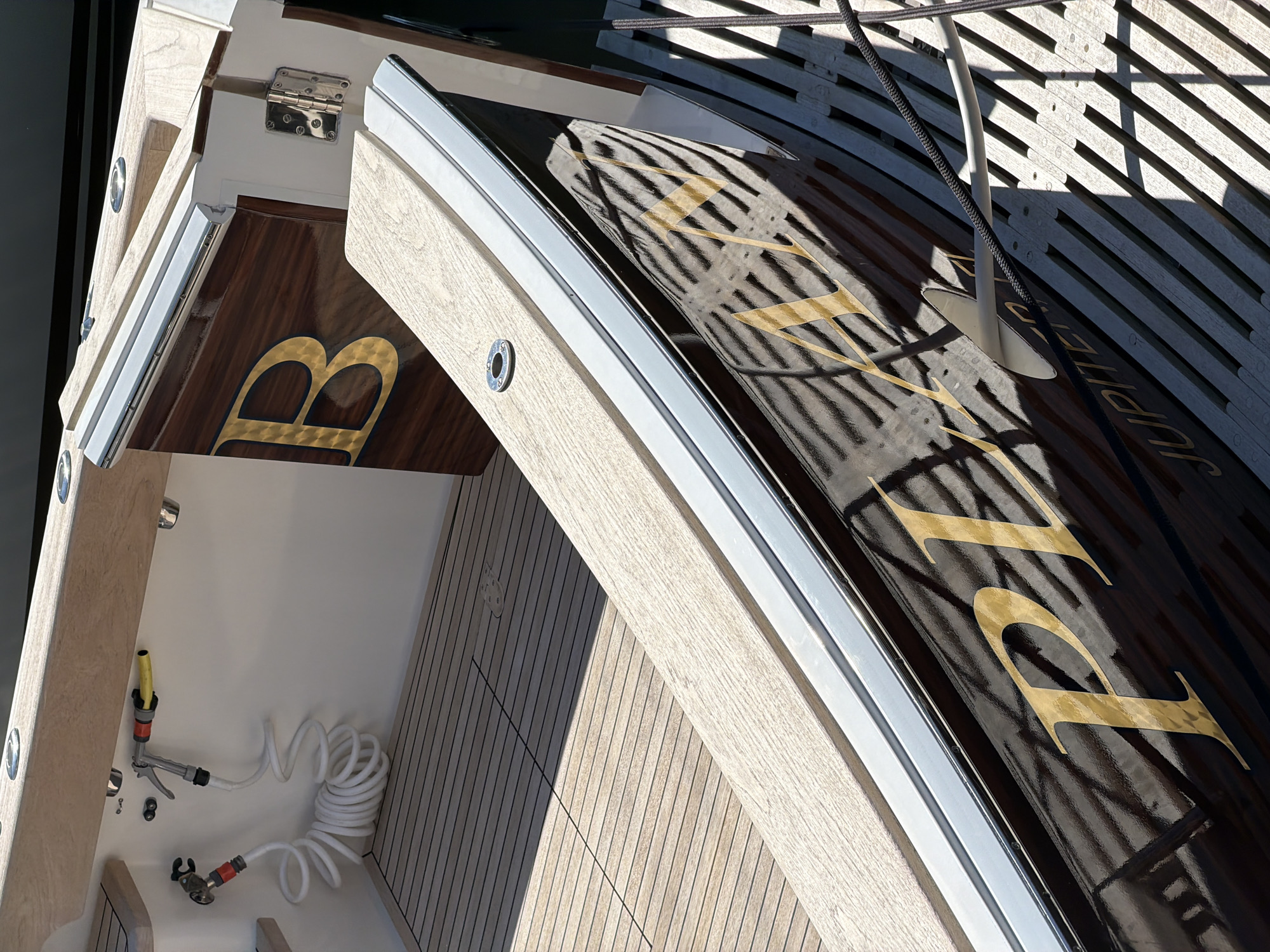 This image shows the stern of a wooden boat docked in the Village of Sag Harbor, United States of America. The boat features a polished wooden deck and a dark, reflective hull with gold lettering that spells out "PLANET" and "JUPITER." A coiled white hose with a nozzle is visible on the port side, attached to the white interior wall of the boat. A stainless steel fitting is visible on the deck. The boat is docked next to a wooden pier. The scene is illuminated by bright sunlight, casting shadows on the deck and hull.