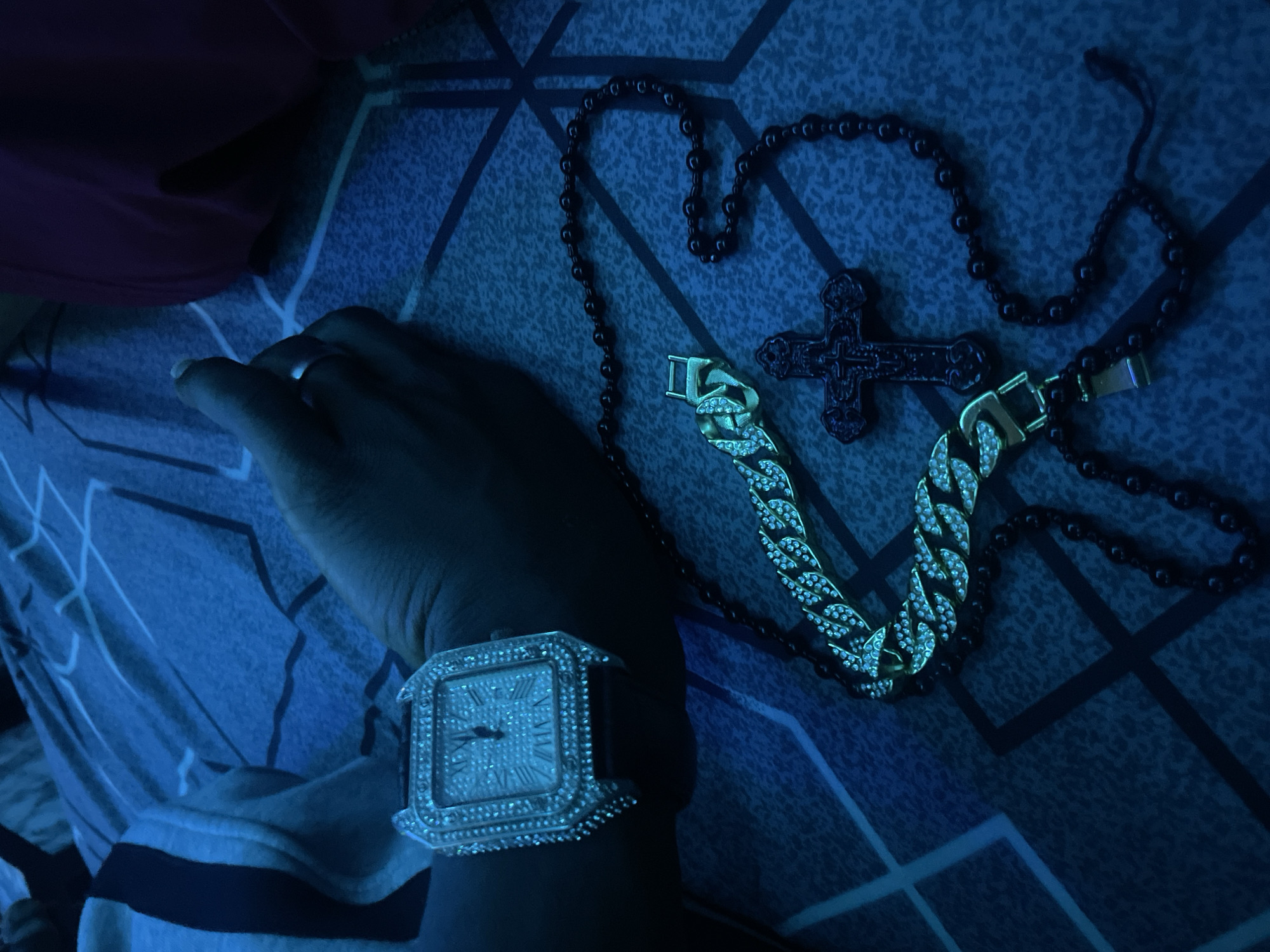 The image shows a close-up of a person's wrist and hand, adorned with a diamond-encrusted watch. To the right of the watch, a cuban link bracelet with diamonds is visible, along with a dark-colored rosary and a crucifix. The objects are arranged on a patterned surface, likely a bedspread or fabric, illuminated by blue lighting. The setting appears to be indoors.