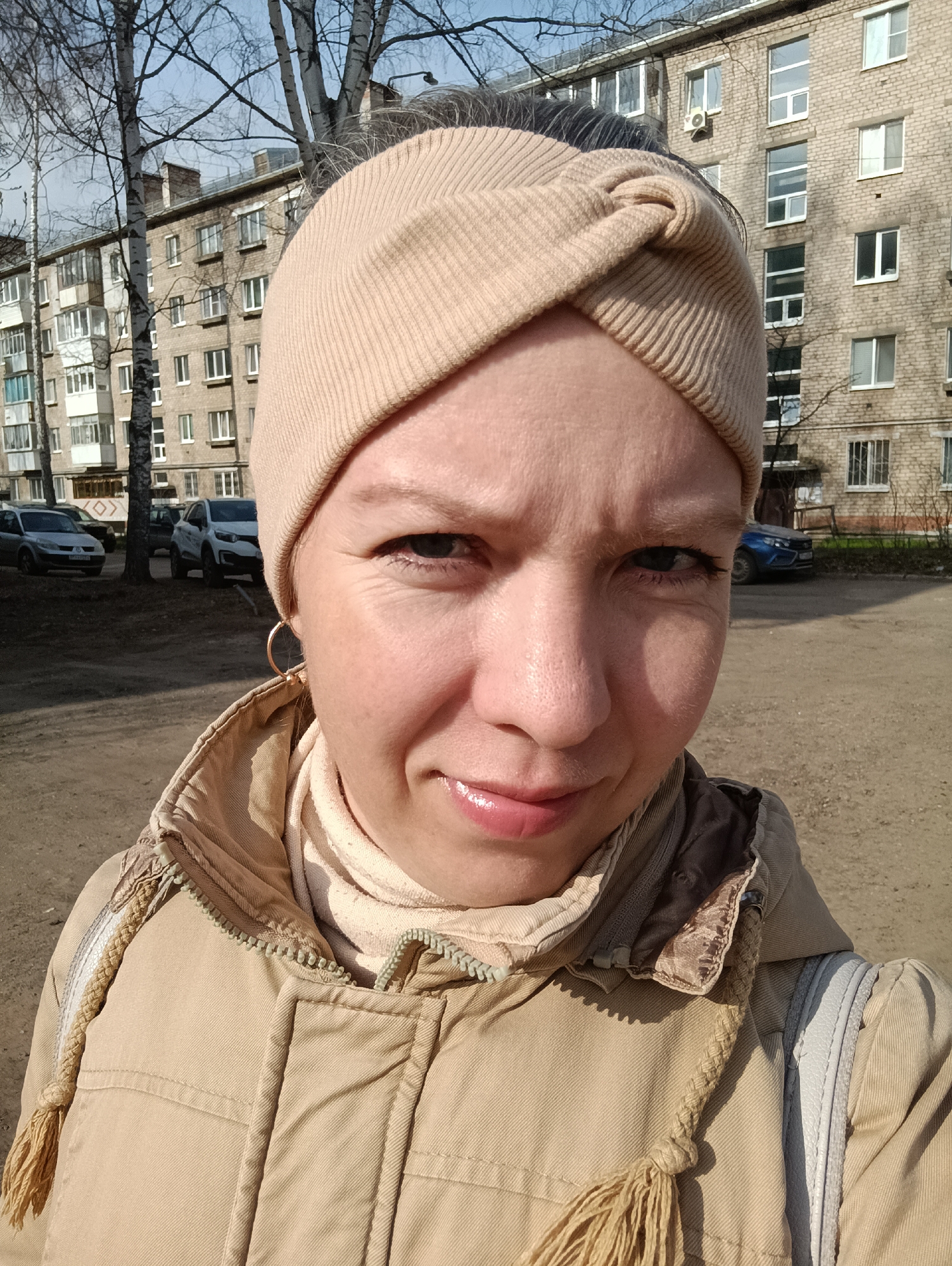 A Caucasian woman is the primary subject of the photograph. She is wearing a light brown headband and a beige-colored jacket with a tassel detail. She has fair skin, grey eyes, and a slight smile. She is wearing a gold earring in her left ear.
The background reveals an urban environment. Several apartment buildings are visible, constructed of tan brick. Cars are parked in the distance. The location is identified as Chaikovsky, Russia, based on the architectural style and the presence of vehicles typical of the region. The ground appears to be dirt or gravel. Bare trees suggest it might be late autumn or early spring. The lighting is bright, suggesting a sunny day. The woman is looking directly at the camera, and the photograph appears to be a selfie.
