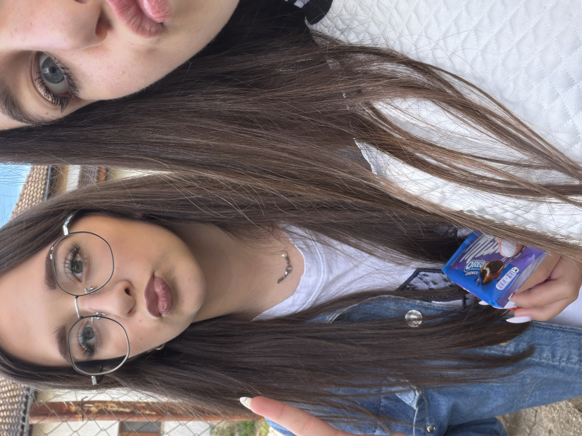 Two young women pose closely together outdoors, holding a small package of Oreo cookies. One woman wears glasses and a denim jacket. The other woman is positioned above, partially out of frame. The location appears to be Zmeitsa, Bulgaria, based on the presence of a structure resembling a fence or wall common in the region.