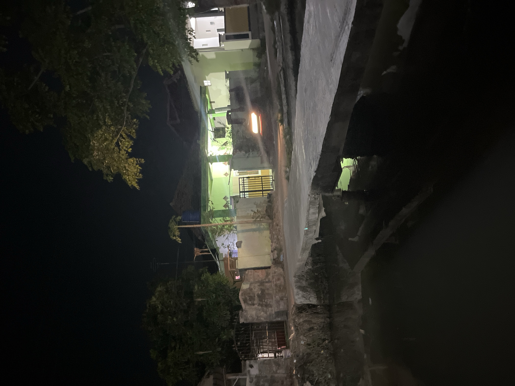 This is a night time image taken in Laban, Indonesia. The scene depicts a rural or suburban area with a body of water, possibly a canal or small river, in the foreground. A concrete bridge spans the water, leading towards a property with a house illuminated by green and white lights. A car with its headlights on is visible on the road near the house, creating a motion blur effect. The property features a gate and a wall. Trees are present in the background, and the overall atmosphere suggests a quiet evening.