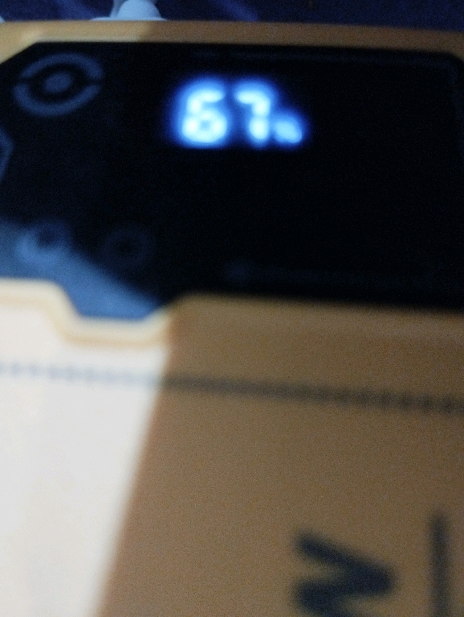 The image shows a close-up of a digital display with blue illuminated numbers. The numbers appear to be "67" with a small dot or comma after the "7". To the left of the display, there is a faint circular or oval shape with concentric rings, possibly a logo or icon. The display is set against a dark background, and a portion of a yellow object is visible in the foreground, out of focus. The environment is not identifiable as being in Kelurahan Sidanegara, Kecamatan Cilacap, Indonesia, or any other specific location.
