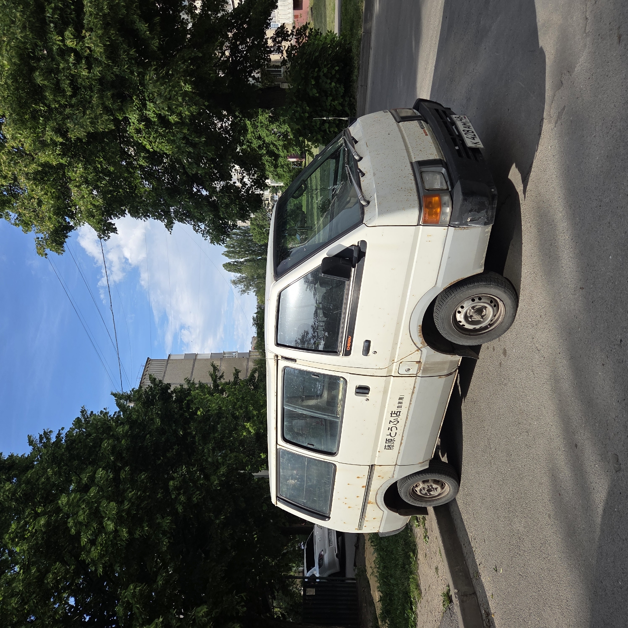 A white, weathered van is parked on the side of an asphalt road next to a concrete curb under a clear blue sky with scattered white clouds. The image is taken in bright daylight, casting distinct shadows on the road, suggesting a sunny day.
The van, which appears to be an older model, possibly a Japanese import given its right-hand drive configuration and side markings, shows significant signs of rust and wear, particularly along its lower body and wheel wells. The front bumper is dark and also shows wear. A license plate, "K 428 AM 48", is visible on the front of the vehicle, indicating a Russian registration (region 48).
Prominently displayed on the side of the van are Japanese characters that read "藤原とうふ店 (自家用)", translating to "Fujiwara Tofu Shop (For private use)", a direct reference to the iconic vehicle from the Japanese manga and anime series "Initial D". Faintly visible on the driver's side window trim is the word "CHERRY".
The van is situated alongside a leafy street, bordered by numerous green trees, some of which form a dense canopy overhead. In the background, a multi-story building with a light-colored facade is partially visible, suggesting an urban or suburban setting. Overhead, thin power lines stretch across the sky. No people are visible in the scene.