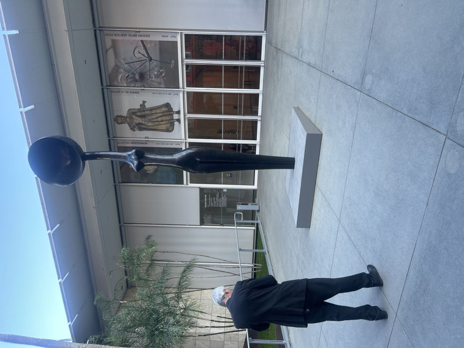 A man in a black suit stands before a dark sculpture of a female form with a large, spoon-like head. The sculpture sits on a gray base in front of a building with glass doors and exhibition posters advertising "Josiah McElheny: Island Universe" and "Realms of the Dharma." Palm trees and a railing are nearby. The background is a modern-style building with a clear sky above. The ground appears to be paved with concrete.