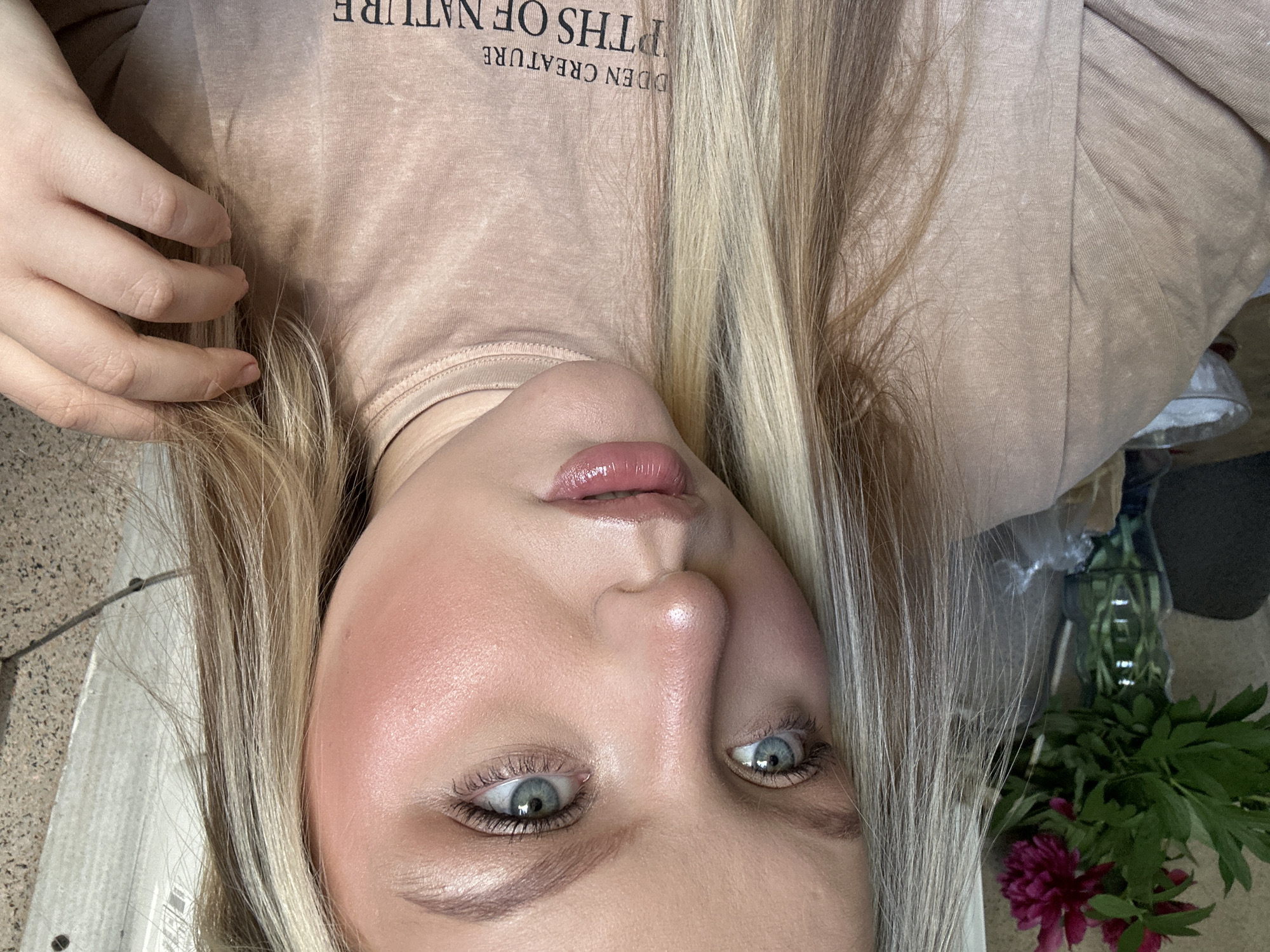 A close-up, selfie-style shot features a blonde woman with blue eyes. She is wearing a beige top with the text "DDEN CREATURE ITHS OF NATURE". Her face exhibits makeup, including blush and lip gloss. Her hand is near her hair. To her left is a floral arrangement with pink flowers in a vase. The setting suggests an interior space, possibly a home. The presence of the floral arrangement and the style of the photograph do not provide enough information to confirm that this image was taken in Chaikovsky, Russia. The location cannot be determined.