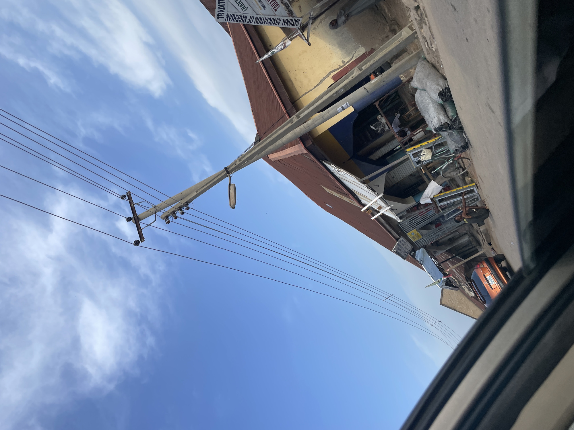 The media file depicts an outdoor scene in Jalingo, Nigeria, likely during the daytime due to the bright blue sky and visible clouds. The perspective is from inside a vehicle, looking outwards and upwards.
The primary subjects are a utility pole with multiple wires stretching across the frame, and buildings lining what appears to be a street or thoroughfare. The buildings have reddish-brown roofs and a yellowish facade, with some sections showing exposed brickwork or concrete. Various objects are visible in front of the buildings, suggesting a marketplace or commercial area. These include shelves stocked with items, sacks of goods, a bicycle, and what looks like a small orange car parked in the distance. A person is partially visible sitting in the foreground of the image, suggesting the photographer is a passenger. Another person is seen in the background near the shelves.
The scene conveys a sense of everyday life in a town or city. The sky is partly cloudy, indicating fair weather. The composition is dominated by the diagonal line of the utility pole and wires against the vast expanse of the sky.
Visible text on a sign above one of the buildings reads "TRADITIONAL" and "NATIONAL ASSOCIATION OF NIGERIAN". This text provides a specific location clue. The overall impression is a busy street scene with overhead infrastructure and commercial activity.