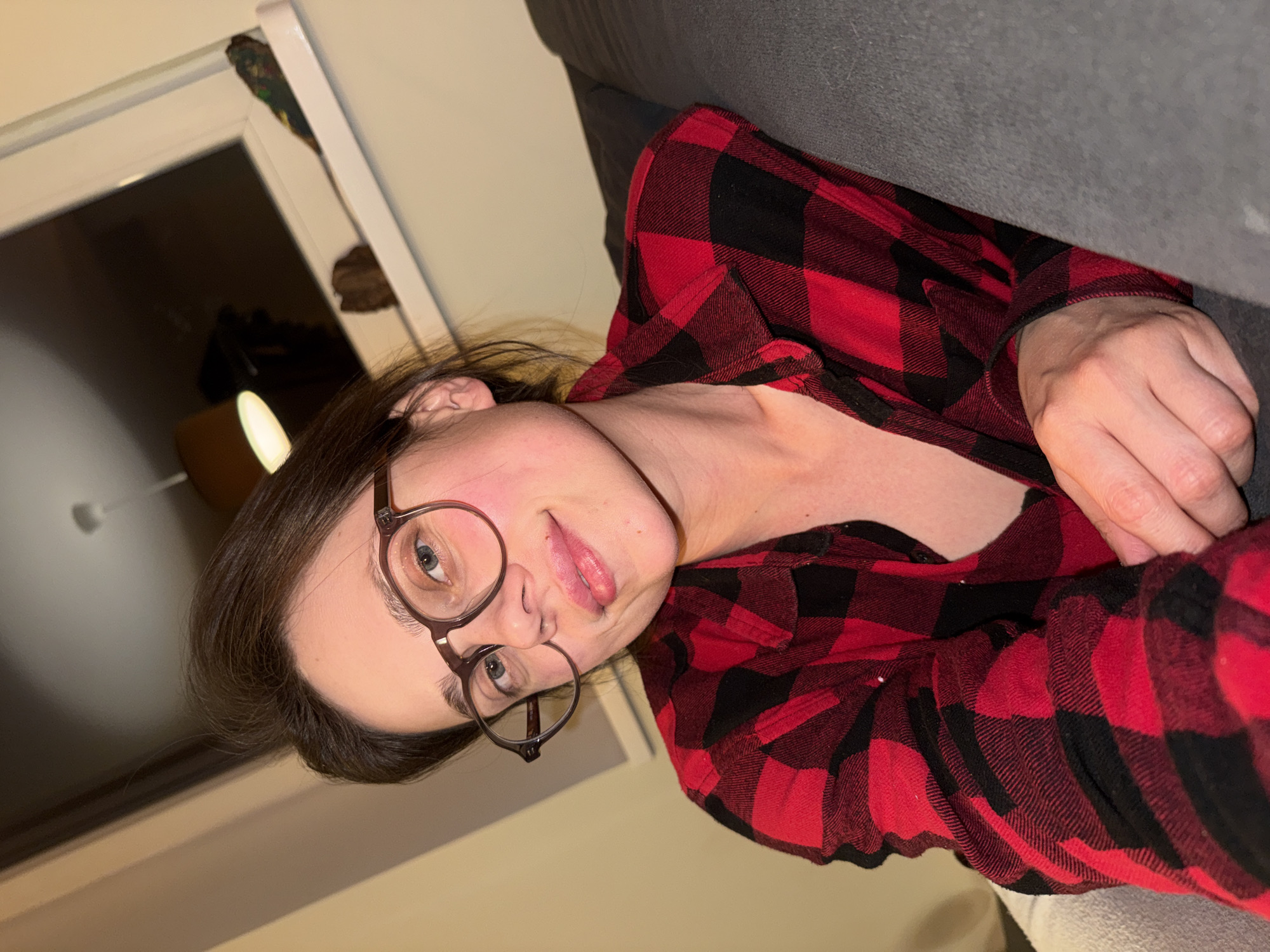 An indoor, casual portrait captures a young person, likely female, in Budapest, Hungary. She is wearing a red and black plaid long-sleeved shirt and round, dark-rimmed glasses. Her dark hair is swept back, and she has fair skin and light blue or grey eyes. She is seen with her head tilted, looking directly at the camera with a playful expression, sticking her tongue out slightly.
The subject appears to be reclining on a dark grey upholstered surface, likely a sofa or couch. One of her hands is visible near her chest/arm area. The background suggests an indoor setting, possibly in the evening or at night, as artificial light illuminates the scene. A white door or window frame is visible to the left, with a dark glass panel, and a hint of a patterned object can be seen tucked into the top of the frame. The overall atmosphere is lighthearted and informal. No discernible text is visible in the image.