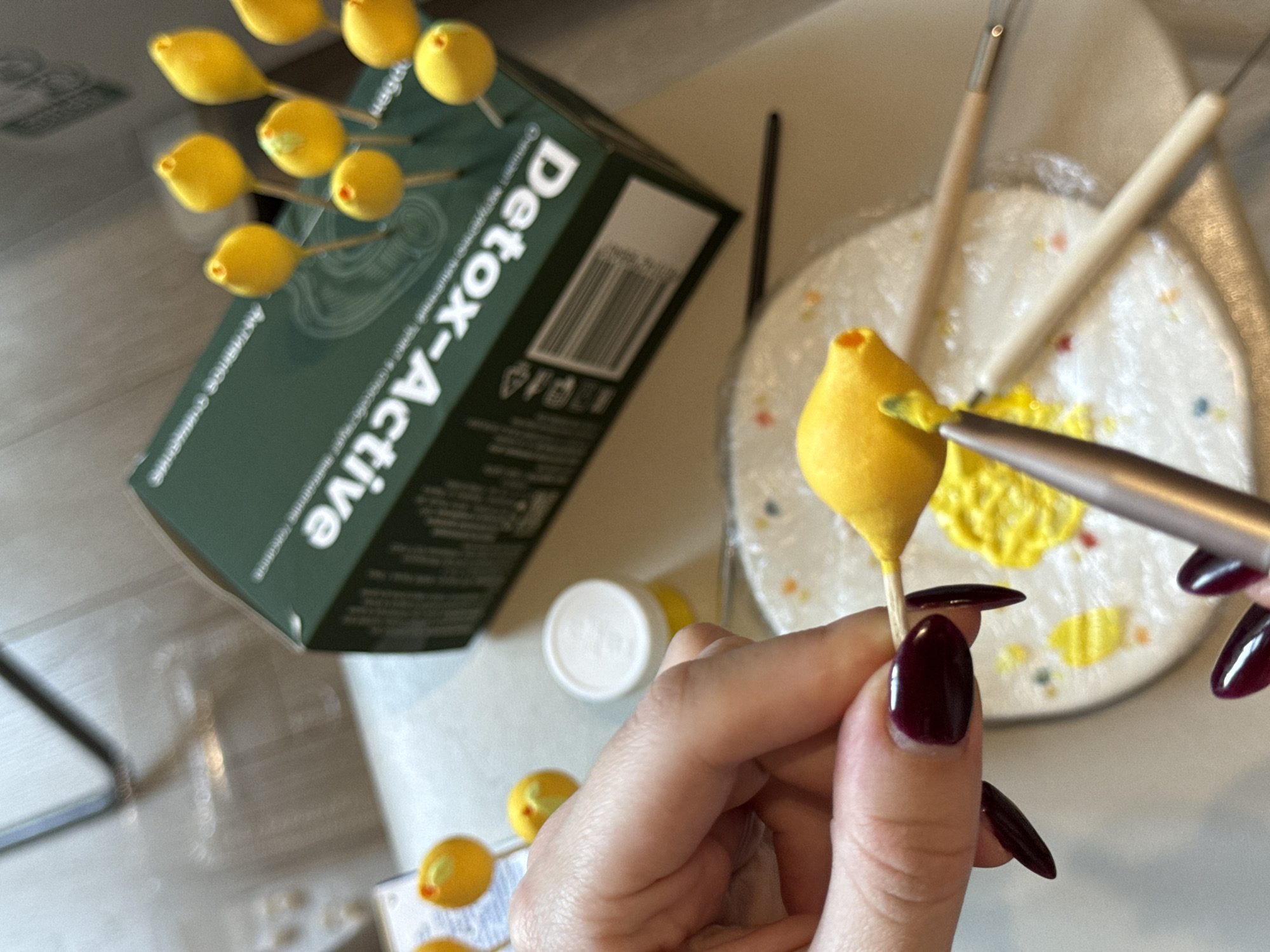 In an indoor environment, a close-up shot focuses on a person's hands as they work on small, yellow, lemon-shaped decorations. The person, with dark red nail polish, is holding one of the decorations with a wooden stick and applying yellow edible paint to it with a tool. Several other finished lemon decorations are visible on sticks, some resting on a white surface with scattered colorful edible sprinkles. A green box with white text, possibly for "Detox-Active," is in the background, along with a small white container of yellow paint. Wooden crafting tools are also present on the white surface.