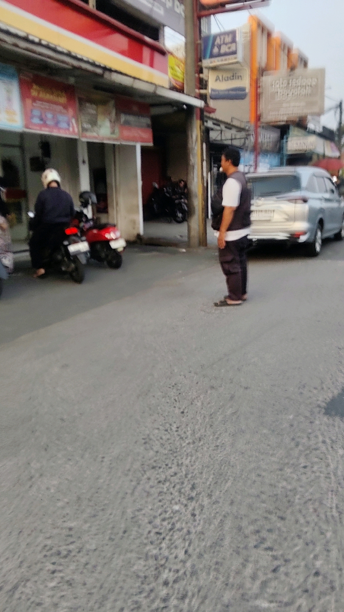 The image depicts a street scene in Pasirsari, Indonesia. In the foreground, the asphalt road surface is visible, with a slight texture. To the right, a man is standing facing away from the camera, wearing a dark vest over a white t-shirt and dark pants. He is wearing sandals. Behind him, a silver car is parked on the right side of the road. In the mid-ground, a red motorcycle is parked, with a person wearing a dark jacket and white helmet seated on it. Another motorcycle is visible behind it. To the left of the road, there is a building with red and yellow signage. Several signs are affixed to the building, including one that reads "ATM BCA" and another that appears to say "Aladin." The scene is likely outdoors, given the lighting and the presence of a street and buildings. The overall impression is one of a typical urban street environment.