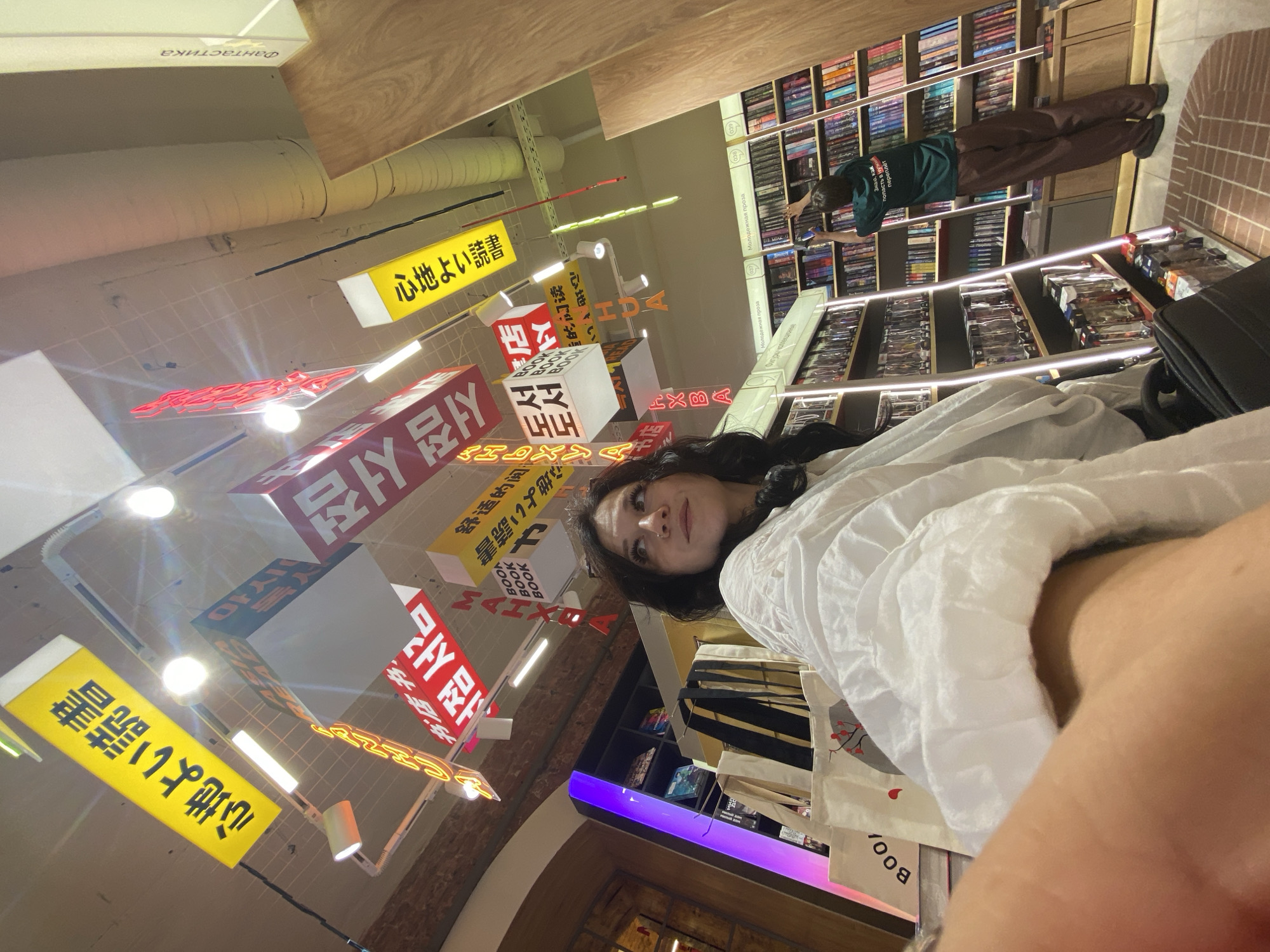 The image depicts the interior of a modern bookstore in Saint Petersburg, Russia. In the foreground, a woman with dark hair, wearing a white puffed-sleeve shirt, is partially visible from the chest up, looking upwards and to the right. Her right arm is extended towards the camera. Behind her, several tote bags are visible, one with the word "BOOK" printed on it.
The ceiling features a display of illuminated signs in various languages, including Japanese and Korean characters, as well as the word "BOOK" in English. These signs hang at different angles, contributing to a vibrant and contemporary atmosphere.
In the background, a person wearing a green t-shirt with text and brown pants is seen reaching for a book on a tall shelf filled with numerous books. This indicates an active bookstore environment with customers browsing. The store features clean, well-lit shelving units and wooden accents. The lighting is a mix of overhead fluorescent lights and the glow from the illuminated signs. The overall impression is that of a well-organized and aesthetically pleasing retail space dedicated to books.