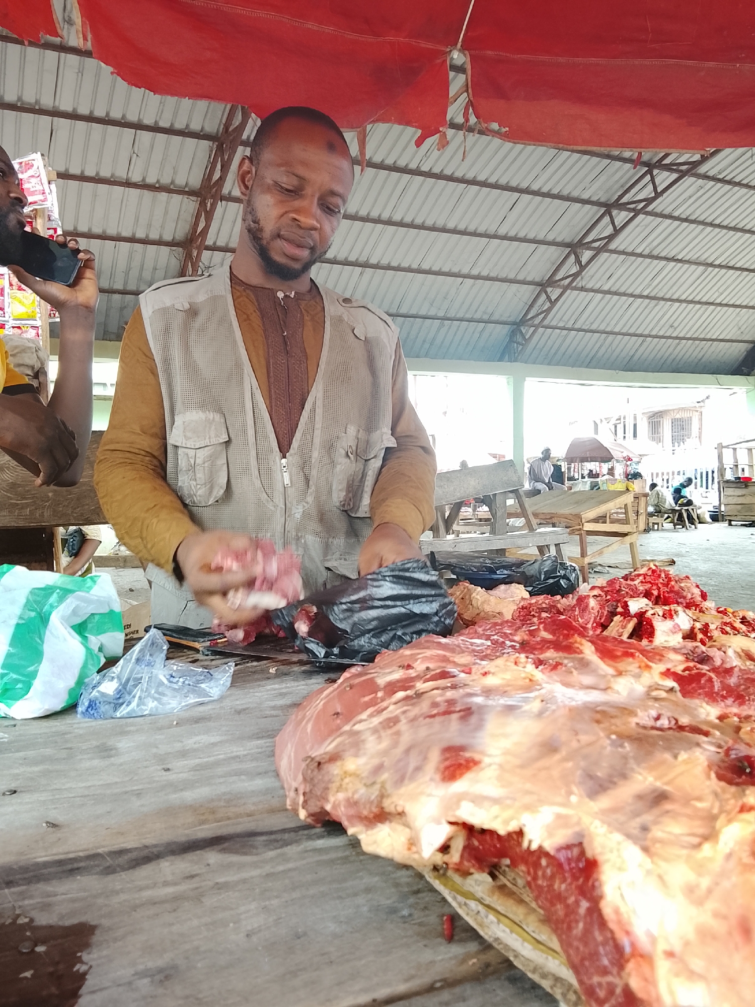 The image depicts a bustling scene within an open-air market in Akwanga, Nigeria, centered around a meat stall. The main subject is a man, likely a butcher, actively engaged in preparing raw meat. He wears a brown long-sleeved shirt under a light-colored, multi-pocketed mesh vest and has a short beard. His expression is focused as he handles pieces of red meat, possibly cutting or portioning them over a black plastic bag or cutting surface on a wooden table.
In the immediate foreground, a large, raw slab of red meat, likely beef, with visible fat and bone, is spread across the worn wooden tabletop. Smaller piles of freshly cut meat are also evident. To the left, a second man's face and arm are visible; he is holding a black smartphone to his ear, seemingly in a conversation. Various plastic bags, including one with green and white stripes, are scattered on the table near the meat.
The setting is covered by a corrugated metal roof supported by metal beams, with a ripped red tarpaulin or canopy hanging below. The background reveals other market activities, with people sitting on wooden benches and indistinct structures under bright daylight. The overall atmosphere suggests a busy, functional marketplace during the daytime. No significant legible text is visible.