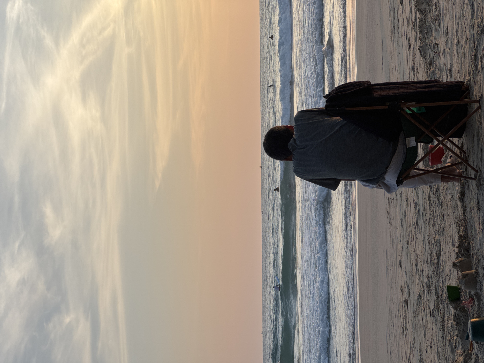 A person sits in a folding chair on the beach facing the ocean at sunset. The person is wearing a grey t-shirt and white pants. A dark towel is draped over the back of the chair. In the foreground, there are children's sand toys, including buckets and sandcastles. The ocean has waves, and several surfers can be seen in the distance. The sky is a soft gradient of orange and pale blue with wispy clouds, indicative of Karon Beach, Thailand.