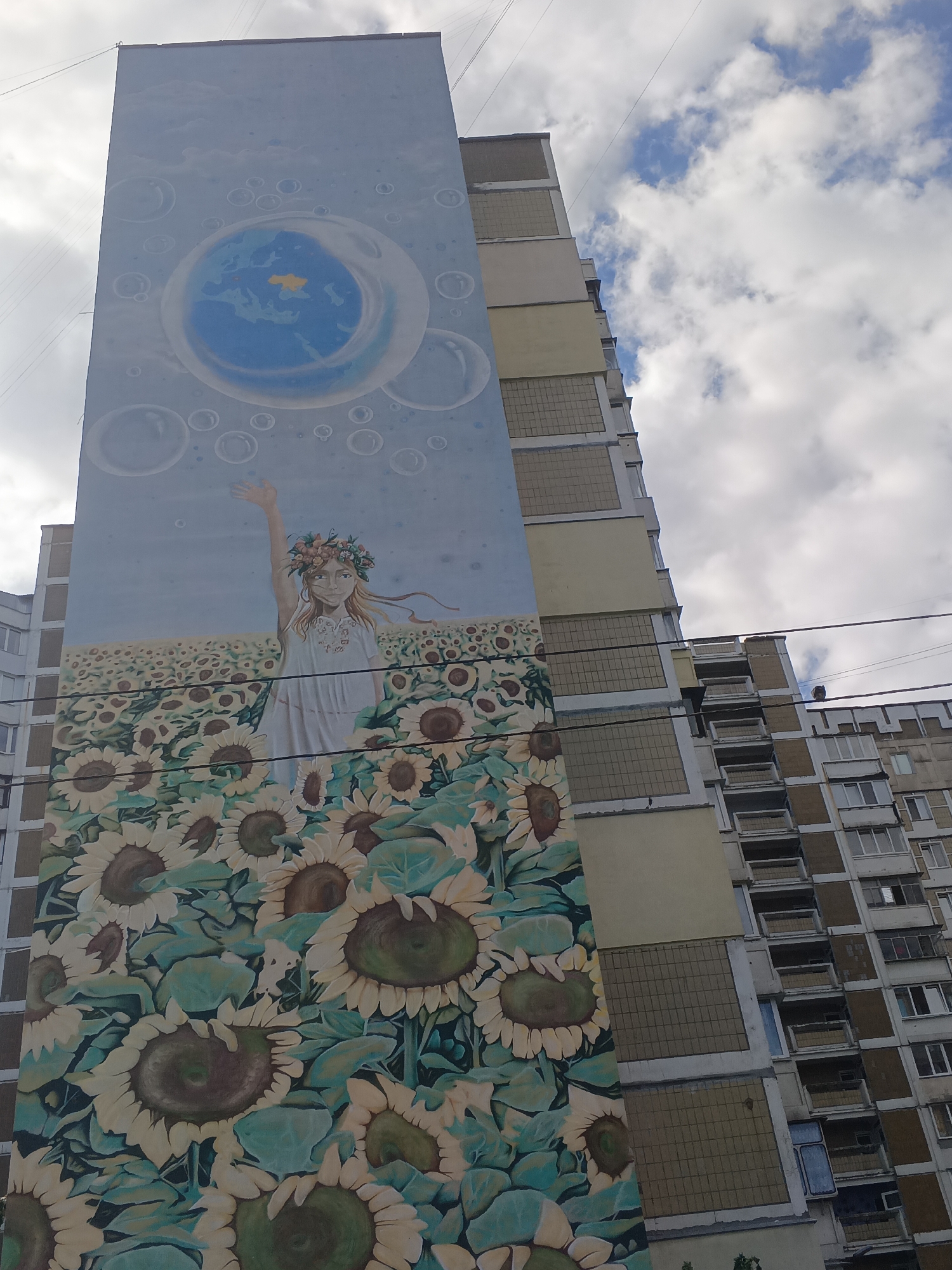 This is a low-angle, vertical photograph of a mural painted on the side of a tall apartment building in Kyiv, Ukraine. The mural depicts a young woman wearing a floral wreath, standing in a field of sunflowers. Above her, the sky is filled with bubbles, and one large bubble contains a depiction of the Earth with Ukraine highlighted in yellow. The building itself is a typical Soviet-era block building, with balconies and windows visible on the right side. The sky is partly cloudy, with white clouds against a blue background. Power lines are visible in the foreground, stretching across the image.