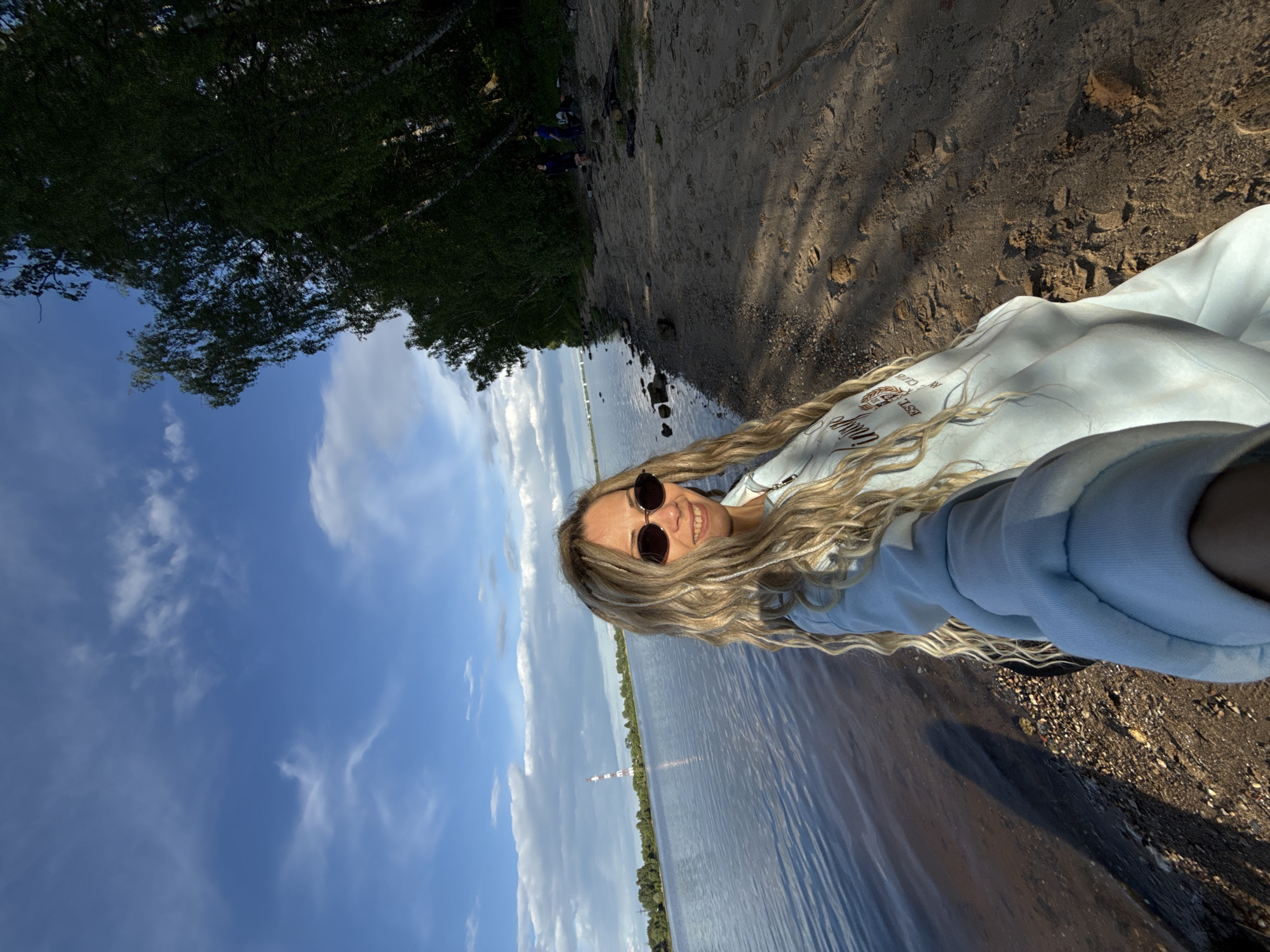 A woman with long, blonde, wavy hair and sunglasses is taking a selfie outdoors. She is wearing a light blue hooded sweatshirt. The background consists of a body of water, trees, and a sky with clouds. A structure resembling a tall chimney is visible in the distance. The terrain underfoot appears to be a mixture of sand and rocks. Based on the location metadata, this image was taken in Шебунино, Russia.