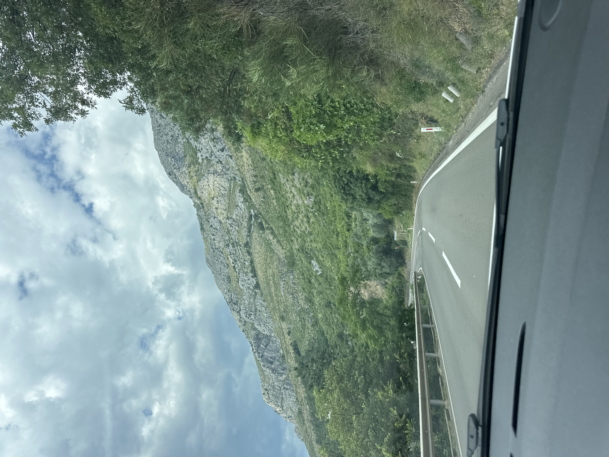 This image captures a scenic drive along a winding road in Rijeka Reževići, Montenegro. The perspective is from inside a vehicle, looking out towards a dramatic mountainous landscape.
The main subjects are the imposing, rocky mountainside covered in lush green vegetation and the asphalt road curving to the right. The road has white lane markings, and a metal guardrail runs along its edge. Trees and bushes line the roadside, with denser foliage on the upper slopes of the mountain. In the distance, the road disappears around a bend, suggesting a journey through a mountainous region. The sky is partly cloudy, with patches of blue sky visible amongst the white and grey clouds.
The scene conveys a sense of travel and exploration, with the natural beauty of the Montenegrin coast being a prominent feature. The time of day appears to be daytime, given the natural light and the visible clouds. The weather seems to be mild, with a mix of sun and clouds. There are no people or specific objects of interest, other than the road infrastructure and the natural elements. No visible text is present in the image.
