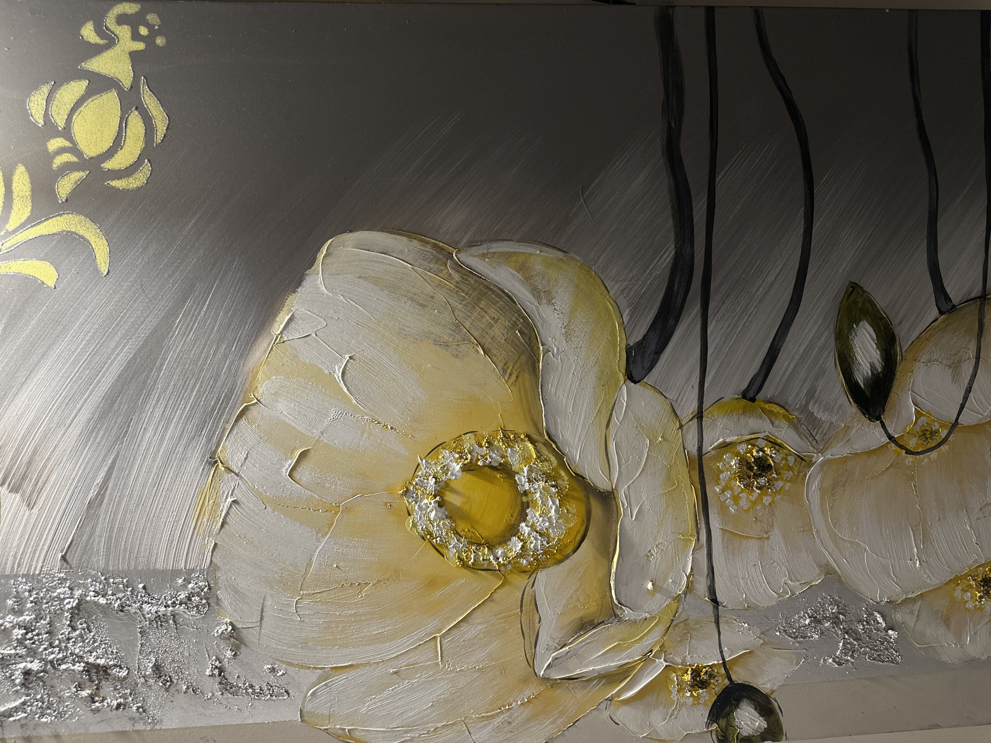 The image displays a close-up of a textured painting featuring large, creamy white and yellow flowers with thick impasto strokes. The centers of the flowers are detailed with yellow and white textured paint, some appearing to have gold accents. Thin, dark stems and leaves are visible beneath the flowers. The background is a brushed silver and gray gradient, with some areas incorporating textured silver elements that resemble distressed metal or foil. Towards the lower right corner, a stylized floral motif in yellow and silver is present.
The painting is located in the city of Peyia, Cyprus.