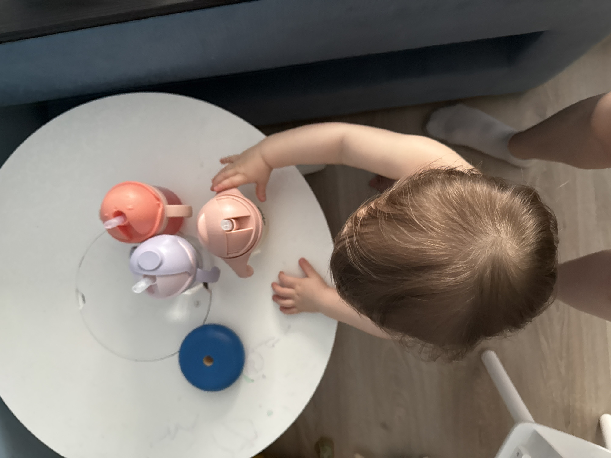 A top-down shot shows a small child at a white, round table with three sippy cups (orange, pink, and lavender) and a blue disc with a hole in the center. The child has light brown hair and is reaching for the pink sippy cup. The child's left foot, wearing a white sock, is visible near the edge of the table, along with part of an adult's leg. Part of a blue sofa is visible in the background. The floor is light brown. The location is not discernible based on the image.