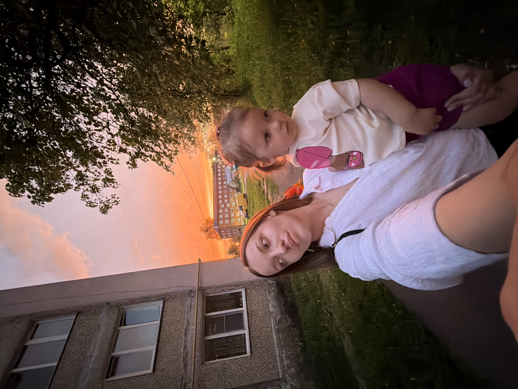 The image captures a selfie of a woman holding a small child, likely a toddler, outdoors in what appears to be Reshetnikovo, Russia. The sunset casts an orange glow across the sky. A gray building with windows is visible in the background. The toddler wears a light-colored shirt and magenta bottoms. The woman wears a white shirt. A tree is visible at the top left. Green grass is visible in the background.