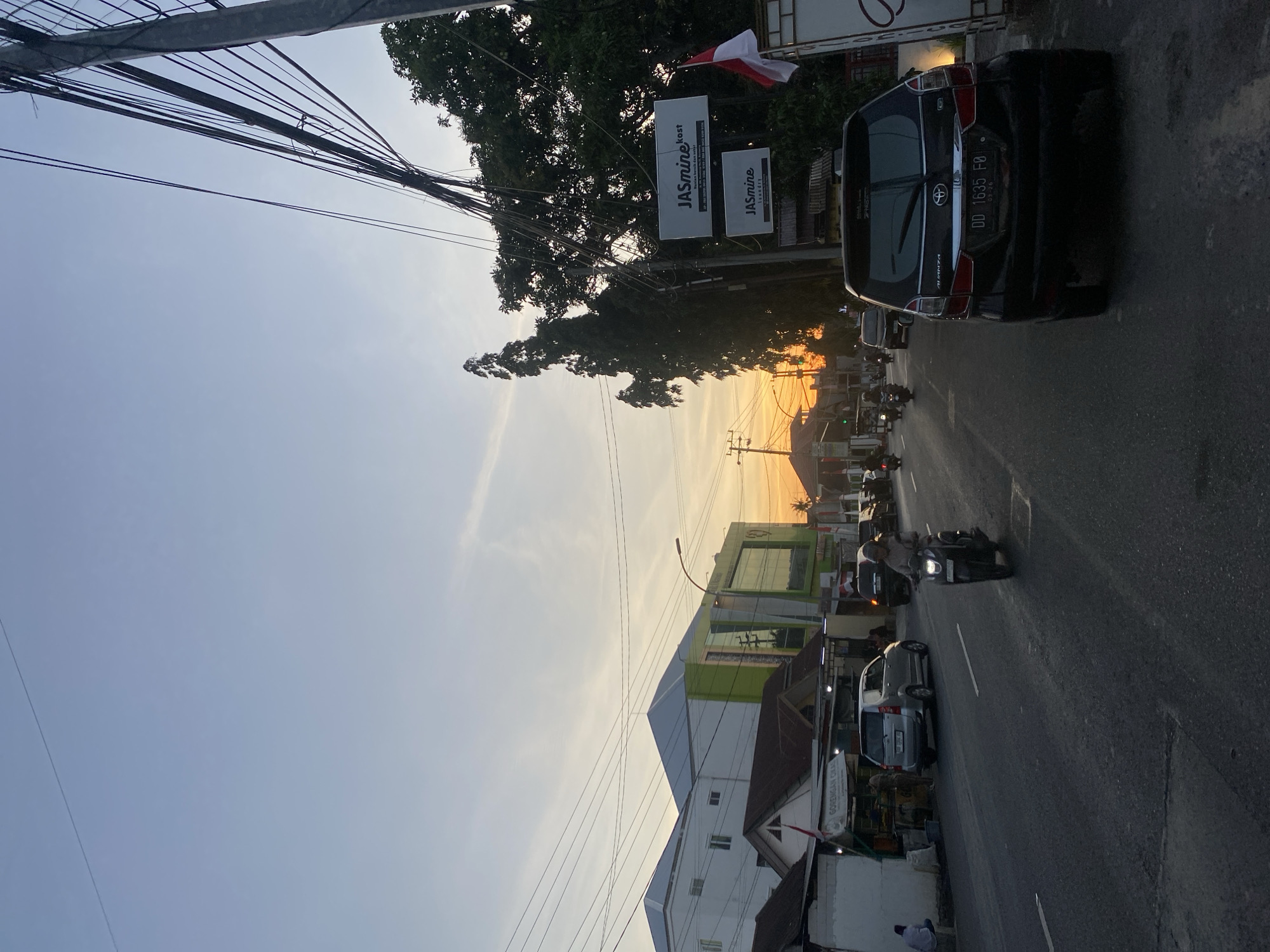 The image depicts a street scene in Bantaeng, Indonesia, during sunset. The sky exhibits a gradient of light blue to orange, indicating the setting sun. A paved road runs through the scene, with several vehicles present. On the left side of the road, a silver minivan is parked, and a person is seated on a low wall. Further down the road, there are buildings, including a white structure with a blue roof and a larger building with green and white accents.
On the right side of the road, a black Toyota Avanza is prominently featured in the foreground, its rear facing the camera. Its license plate reads "DD 1635 F8". Behind the Avanza, a motorcycle is visible, with its rider wearing a helmet and light-colored clothing. Several other motorcycles are also on the road, moving in the same direction as the Avanza.
A utility pole with numerous wires is situated on the far right of the image, partially obscuring some of the trees and buildings. Two signs are attached to the utility pole. The upper sign says "Jasmine kost" and the lower sign says "Jasmine laundry". A small Indonesian flag is also attached to the pole. Dense green trees are visible behind the utility pole. The overall impression is of a bustling urban environment at dusk, with the warm colors of the sunset casting a glow over the scene.