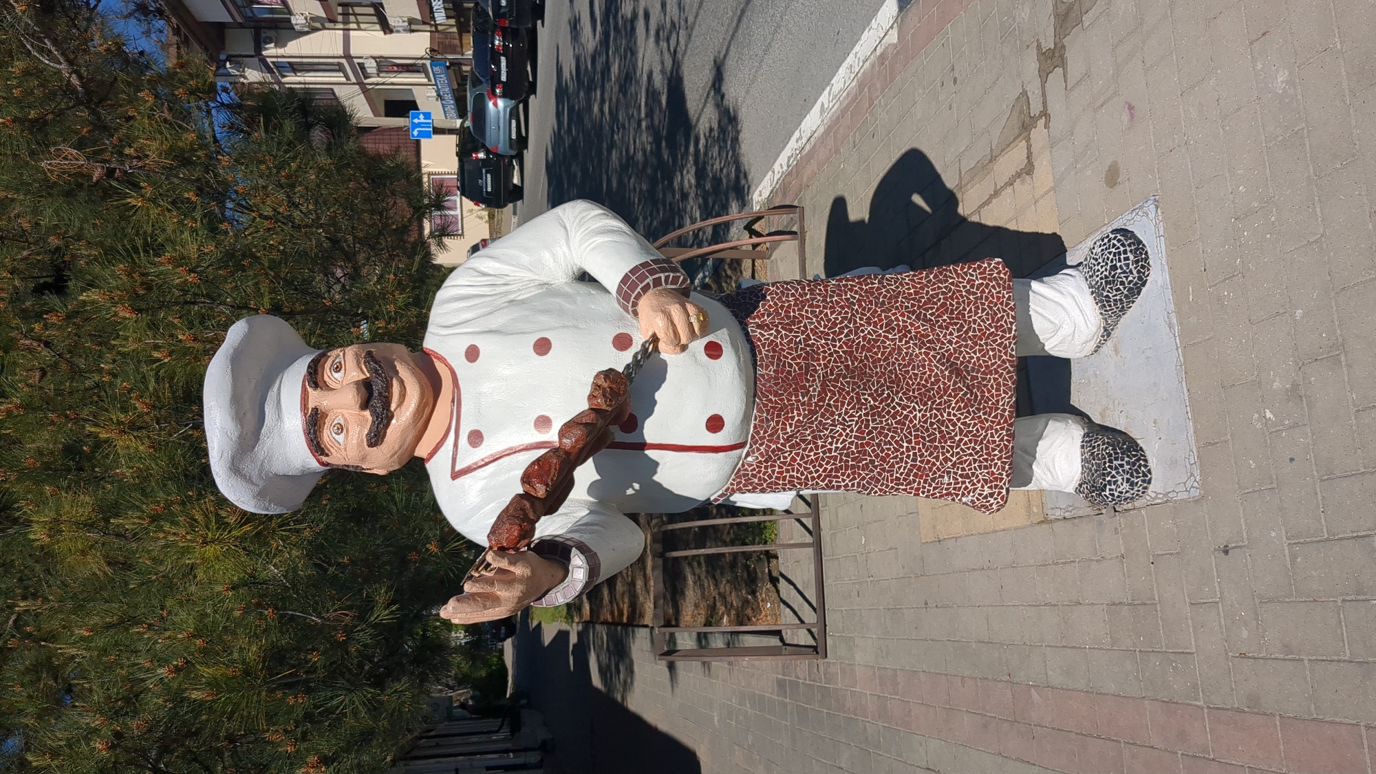 A large, colorful statue of a chef stands on a paved sidewalk, depicting a jovial character holding a skewer of grilled meat (shashlik). The chef wears a white chef's hat, a white coat with red polka dots, a brown and white patterned apron, and dark patterned shoes. A gold ring is visible on one of his fingers. The statue has a friendly expression with a prominent mustache.
Behind the statue, on the left, are dense green pine trees, and across a street, parked cars line the road in front of multi-story buildings with balconies. The scene is brightly lit by the sun, casting long shadows on the sidewalk, suggesting it is either morning or late afternoon. The sky appears clear.
On a blue sign in the background, visible above the parked cars, Cyrillic text can be seen, which reads "СТОМАТОЛОГИЯ" (Dentistry), along with a blue arrow sign below it. The statue stands on a light-colored rectangular base. The overall setting appears to be an urban or suburban commercial area in Gelendzhik, Russia.