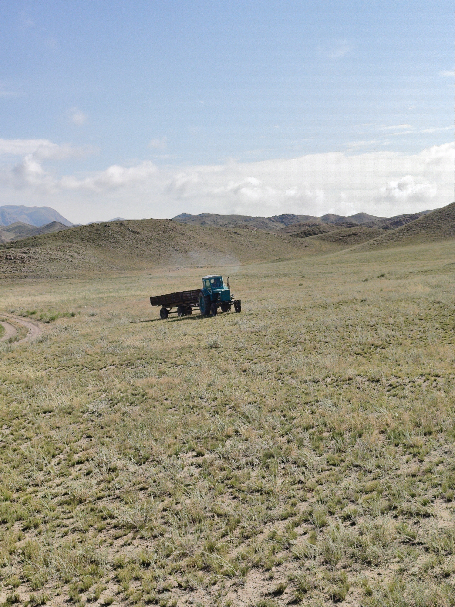 A teal tractor with a dark brown trailer is moving across a dry, grassy plain. The tractor is emitting a faint plume of blue exhaust. The terrain is gently rolling with sparse vegetation. In the background, there are several hills or mountains, some with rocky outcrops. The sky is a light blue with scattered white clouds. A dirt road is visible in the lower left corner of the image.