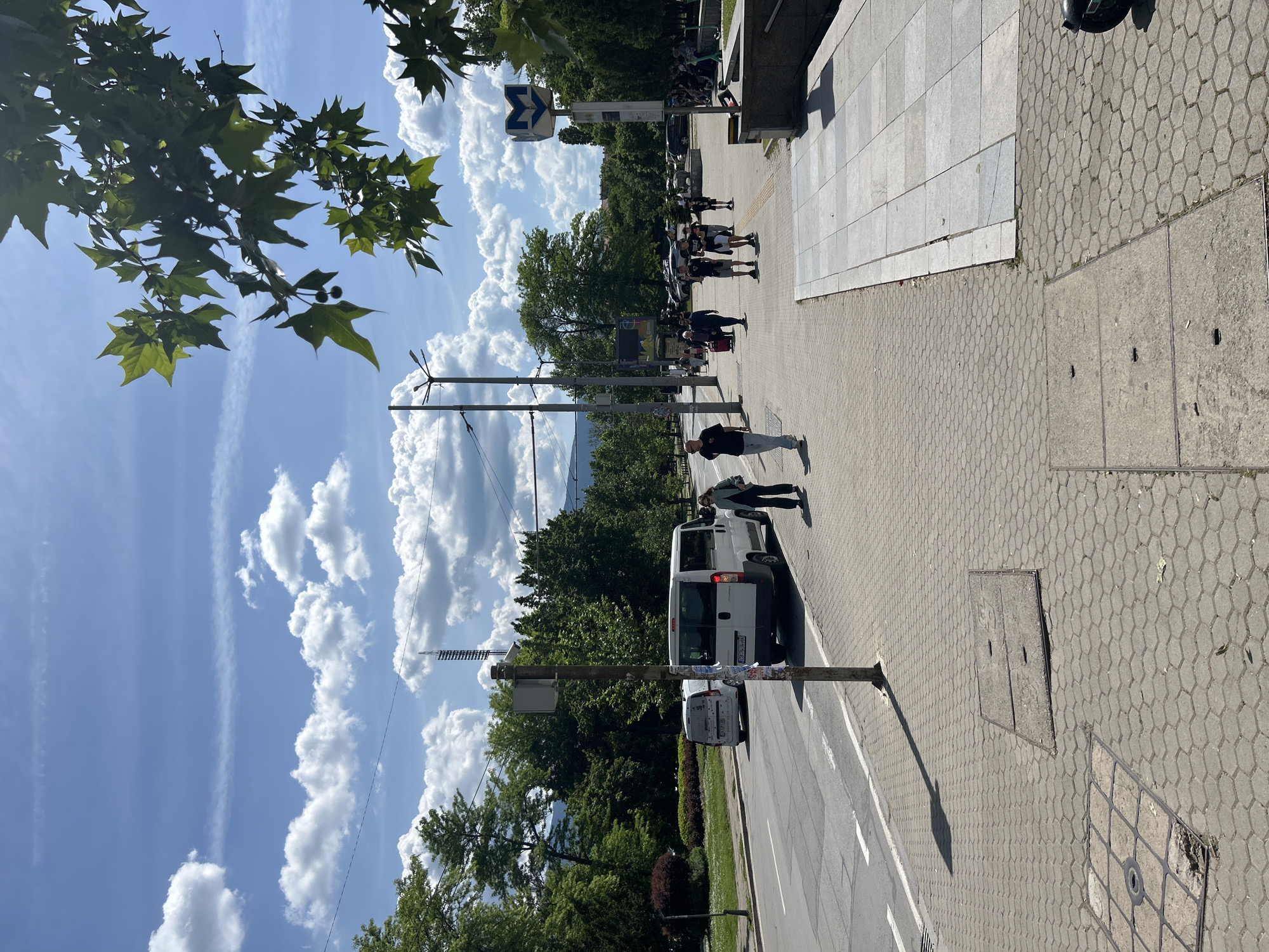 A street scene is captured in this image, showing a pedestrian walkway, street with vehicles, and a partly cloudy sky. The location is identified by a blue-and-white metro sign, indicating the presence of a metro station in the vicinity. People are observed walking on the pedestrian walkway, suggesting the area is accessible to the public. A white van is parked on the side of the road, and other vehicles can be seen in the distance. The environment includes trees, which line the street and add greenery to the scene. The paving stones on the walkway exhibit a honeycomb pattern. Utility poles are present, and the sky features contrails.