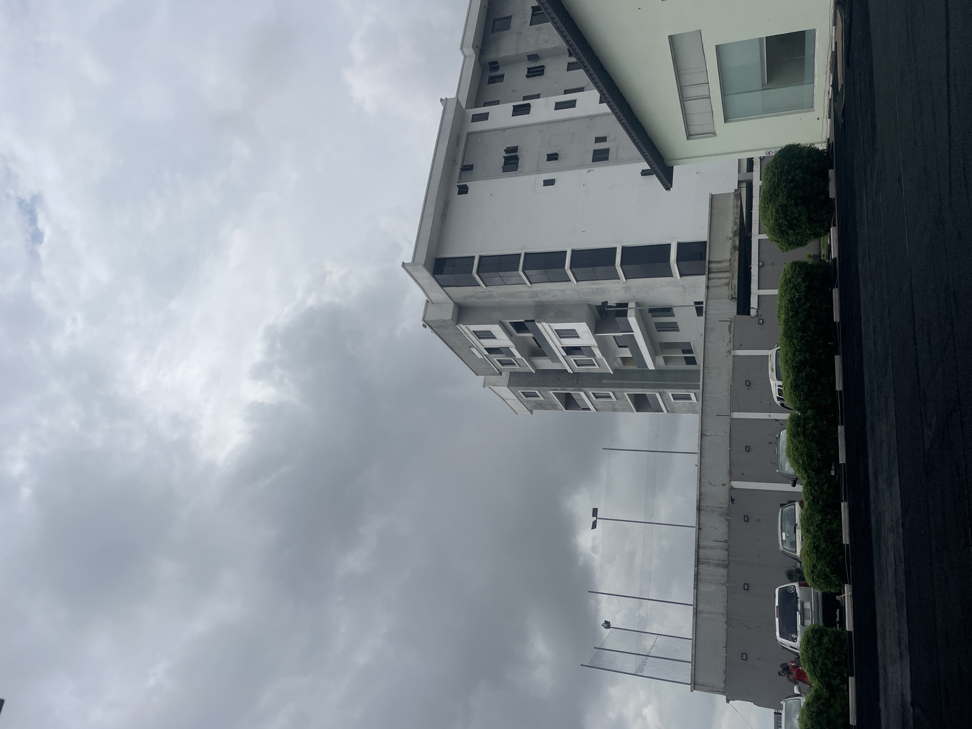 The image depicts an overcast sky above a modern residential building in Moba, Nigeria. The building, which appears to be under construction or recently completed, is multi-story with a combination of white and grey facades and dark-tinted windows. In the foreground, a line of neatly trimmed green bushes separates a dark asphalt area from a row of parked cars, including SUVs and sedans. A concrete wall with a chain-link fence extends across the midground. The overall scene suggests an urban or suburban environment, with the presence of residential architecture, vehicles, and public amenities.