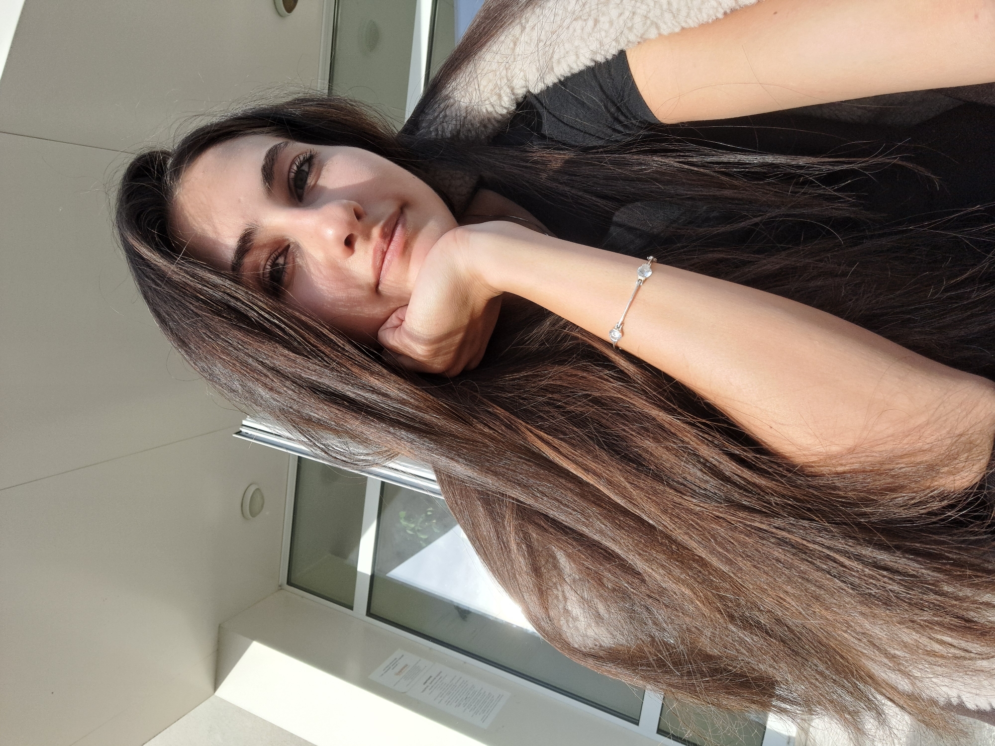 A woman with long, brown hair is resting her head on her hand, which is adorned with a silver bracelet featuring two clear stones. She is wearing a black shirt and a light-colored jacket. A window is visible in the background. Based on the location, it's presumed to be in or near Chepintsi, Bulgaria. The photo appears to be taken indoors, with natural lighting coming through the window.