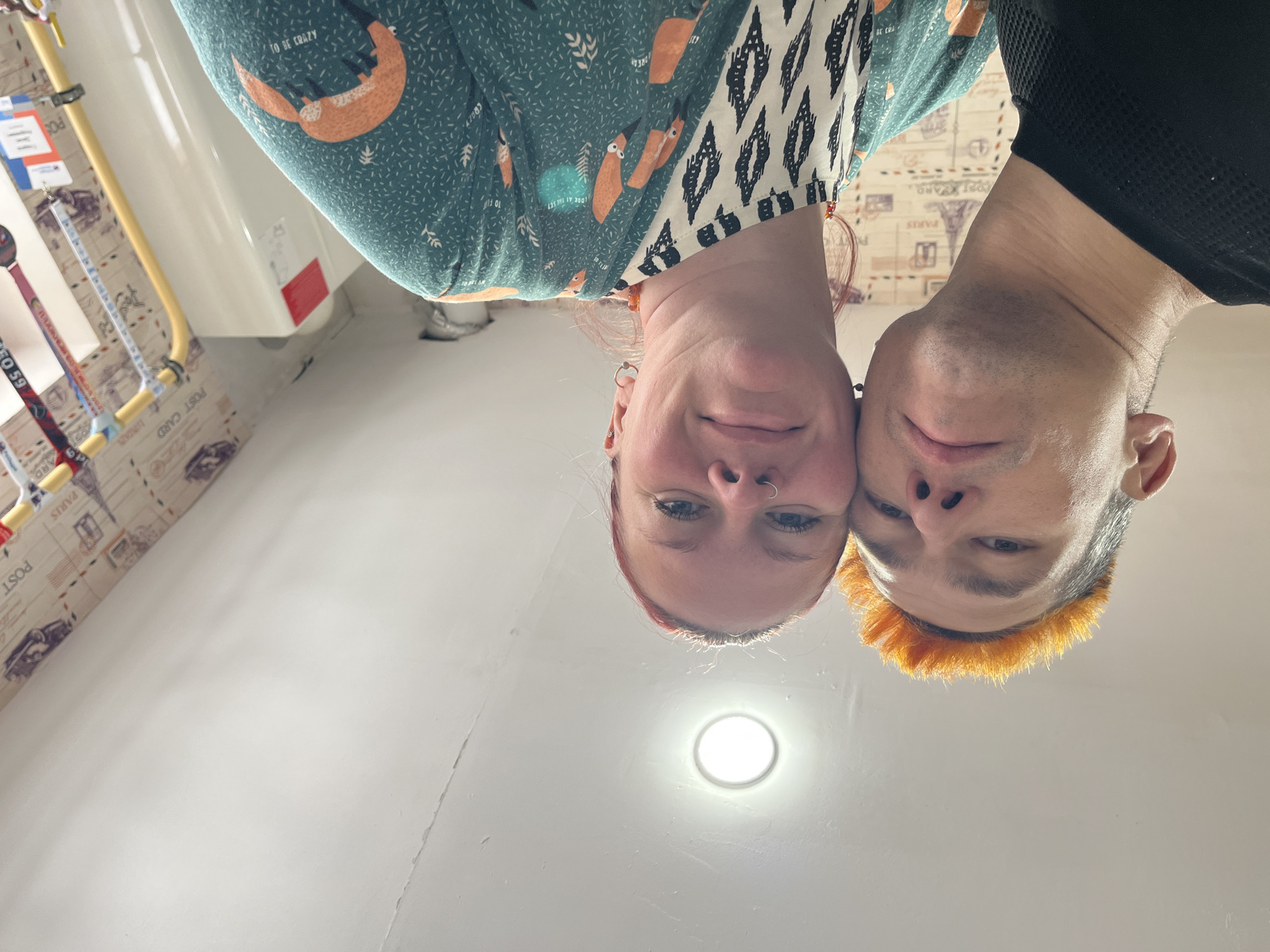 The image is an upside-down, close-up shot of two people. One person has red hair and a nose ring, wearing a green shirt with fox patterns and black and white designs around the collar. The other person has short, orange hair and wears a black shirt. They are close together, almost touching heads. The background shows a white ceiling with a circular light fixture. The wallpaper has a "Paris" theme with postcard images and text. There are pipes and other items attached to the wall. The image does not appear to be located in Chaikovsky, Russia.