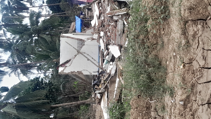 A destroyed building stands amidst rubble, likely caused by an earthquake. Remnants of brick and white walls are visible. The ground is dry and cracked, with sparse vegetation. Palm trees line the left side of the frame. In the background, other buildings can be seen along with what appears to be a blue tarp. The scene suggests devastation and destruction.