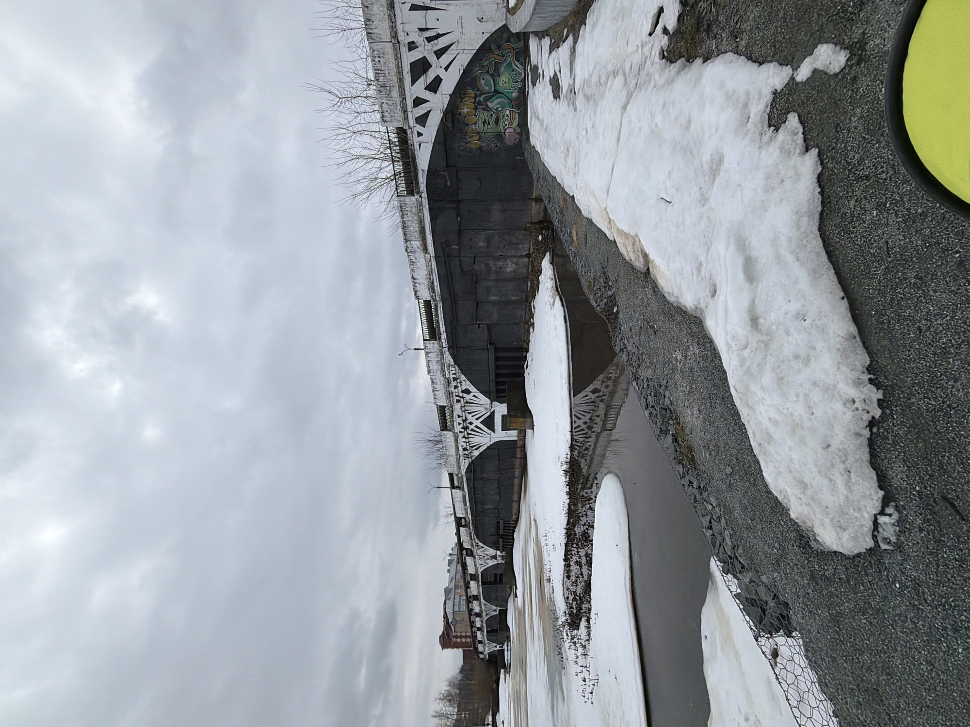 The image shows a partial view of the bridge crossing the Votka River in Votkinsk, Russia. The sky is overcast and gray. Patches of snow are present along the riverbank, which is covered in gravel and reinforced with wire mesh. The bridge's white structure features decorative geometric patterns and some graffiti on the supporting pillars. The river reflects the bridge's arches and the cloudy sky. Trees without leaves can be seen in the distance. A portion of a yellow object appears in the upper right corner of the image.