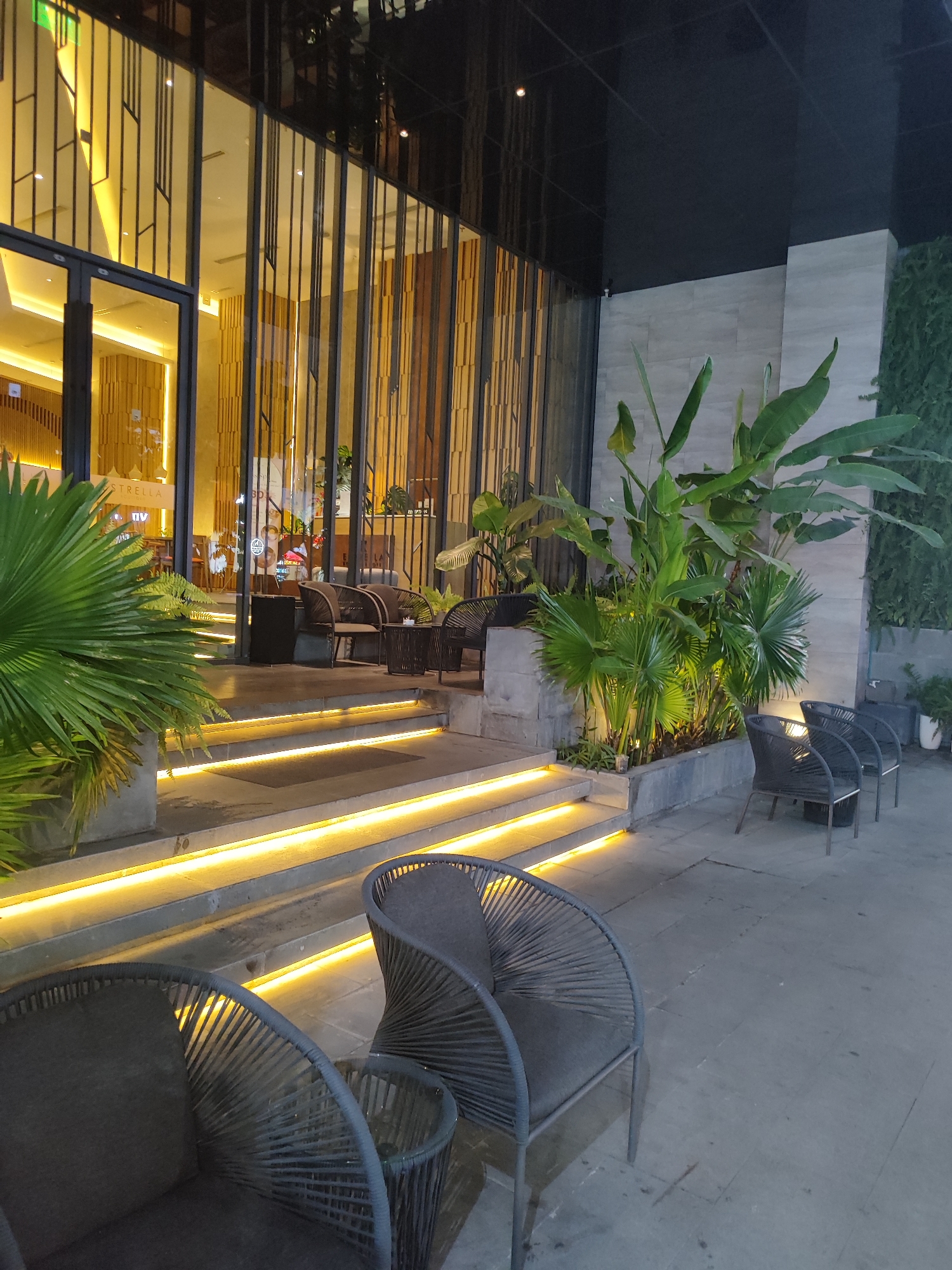 This media file depicts the exterior of a modern establishment, likely a cafe or restaurant, at night in Da Nang, Vietnam. The scene is illuminated by warm, ambient lighting, with bright yellow LED strips accentuating a set of outdoor stairs. The architecture features a glass facade with vertical metal accents, reflecting the interior lights.
In the foreground, several contemporary outdoor chairs with dark upholstery and woven detailing are arranged on a paved patio. One chair is prominent in the lower left, with another to its right, accompanied by a small glass-topped table. Further back, more chairs and tables are visible on a raised platform leading into the building, suggesting an outdoor seating area.
Lush green plants, including large-leafed tropical varieties and smaller palm-like plants, are strategically placed around the entrance, adding a touch of nature to the urban setting. The greenery is also lit, creating a soft glow.
The interior of the establishment is visible through the glass, showing a warmly lit space with wooden paneling and what appears to be a sign with the word "ESTRELLA" on it. The overall impression is one of a sophisticated and inviting nighttime ambiance. The weather appears to be clear, given the nighttime setting and the absence of rain. No people are visible in the media, suggesting the establishment may be closed or patrons are inside. The lighting and the presence of outdoor seating indicate this is a commercial space designed for customer enjoyment.