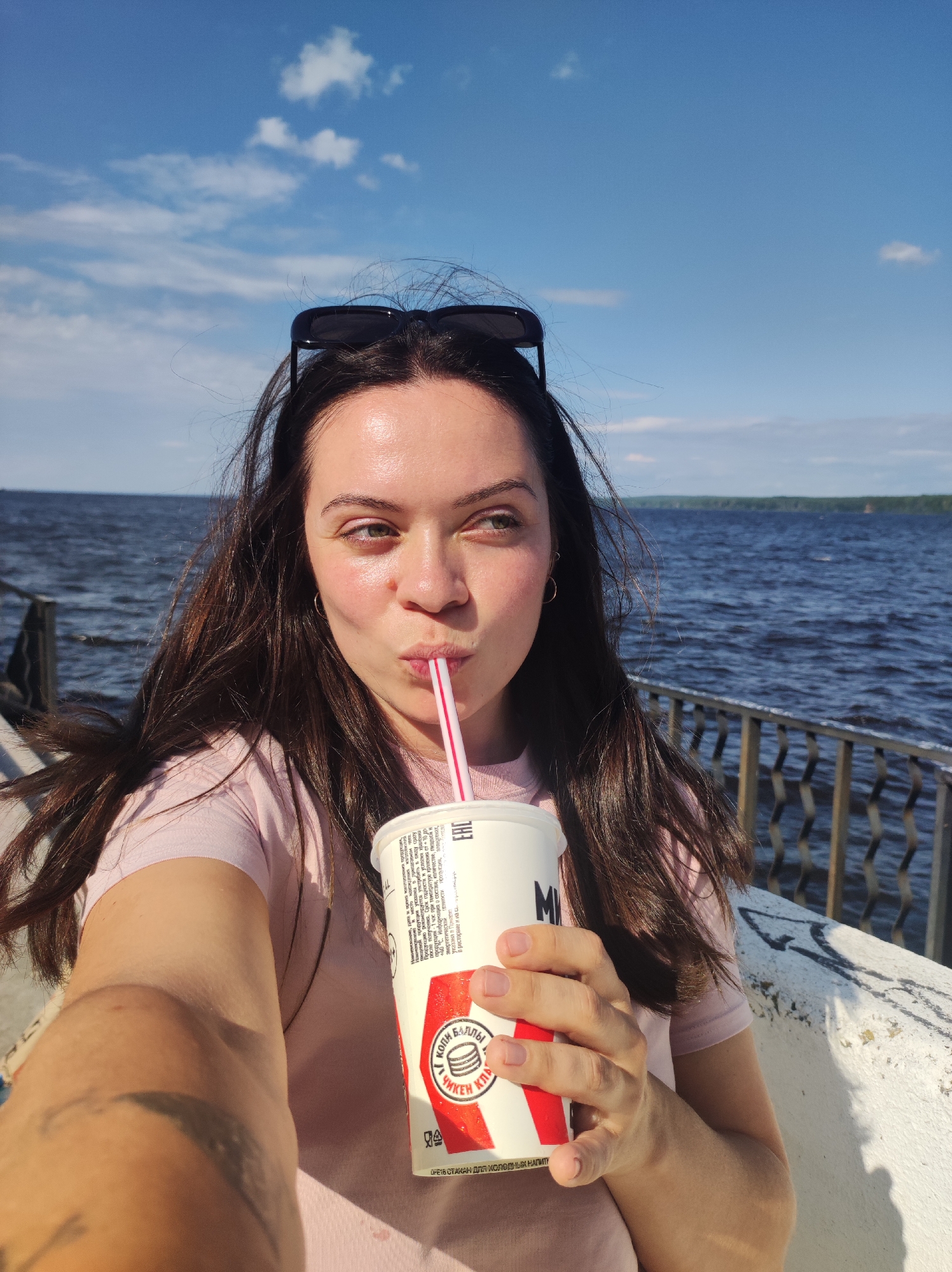 A young woman with long, dark hair is drinking from a large white cup with a red and white design. She is wearing a pink t-shirt and sunglasses are resting on top of her head. She is holding the cup with her right hand and a pink and white straw is in her mouth. The woman is looking off to the right side of the frame with her lips pursed. Behind her, a body of water stretches to the horizon, with a line of trees visible in the distance. A metal fence runs along the edge of the water. The sky is blue with scattered white clouds, suggesting it is a sunny day. The text on the cup is partially visible and appears to be in Russian. The scene suggests a casual outing by the water on a pleasant day.