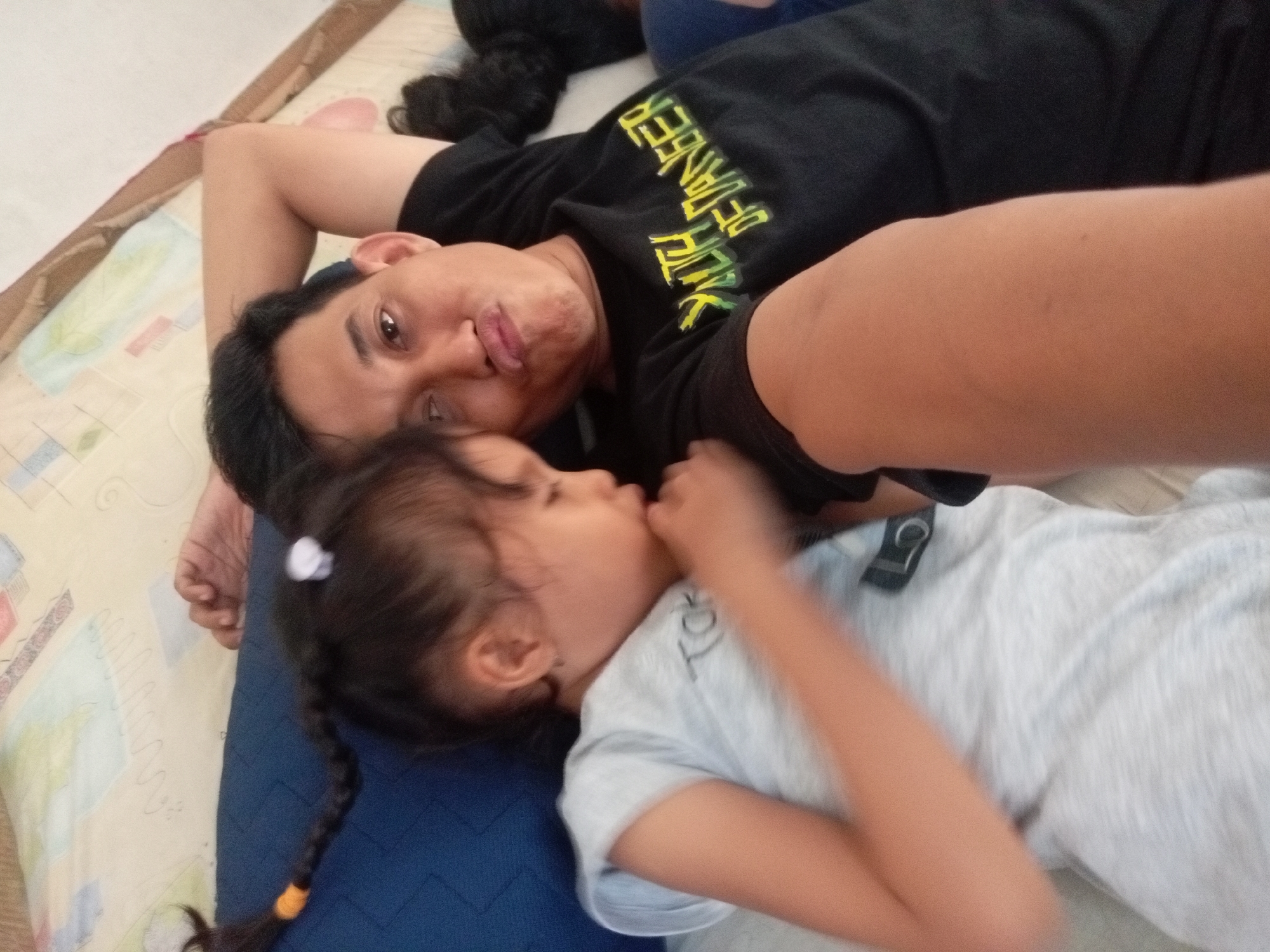 A father and daughter are lying on a blue mattress, with the father on top. The father is wearing a black t-shirt with yellow lettering and the daughter is wearing a light gray t-shirt. The father is looking up at the camera, while the daughter is looking down. They are in a bedroom, likely in a home. The background is blurred, but it appears to be a bed with a patterned sheet. This image was taken in Batam, Indonesia.