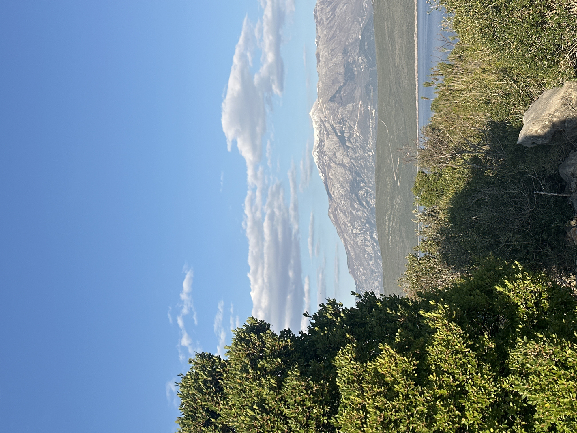 The image shows a landscape in Glavaticici, Montenegro. In the foreground, there is green shrubbery. Behind this, a mountain range is visible, with some snow on the peaks. A thin strip of water separates the mountains from the shrubbery. Above this, there is a clear blue sky with several fluffy white clouds.