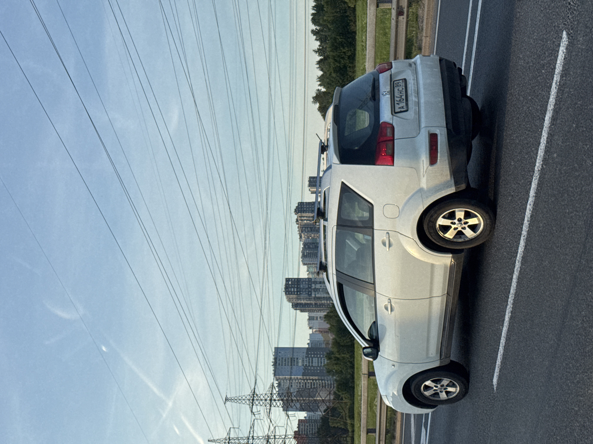 A white Peugeot 4007 is driving on a highway. The car is a crossover SUV, with a roof rack and tinted windows. The license plate reads "A 164 HC 89". In the background, there are tall apartment buildings, power lines, and trees. The sky is blue with some clouds.