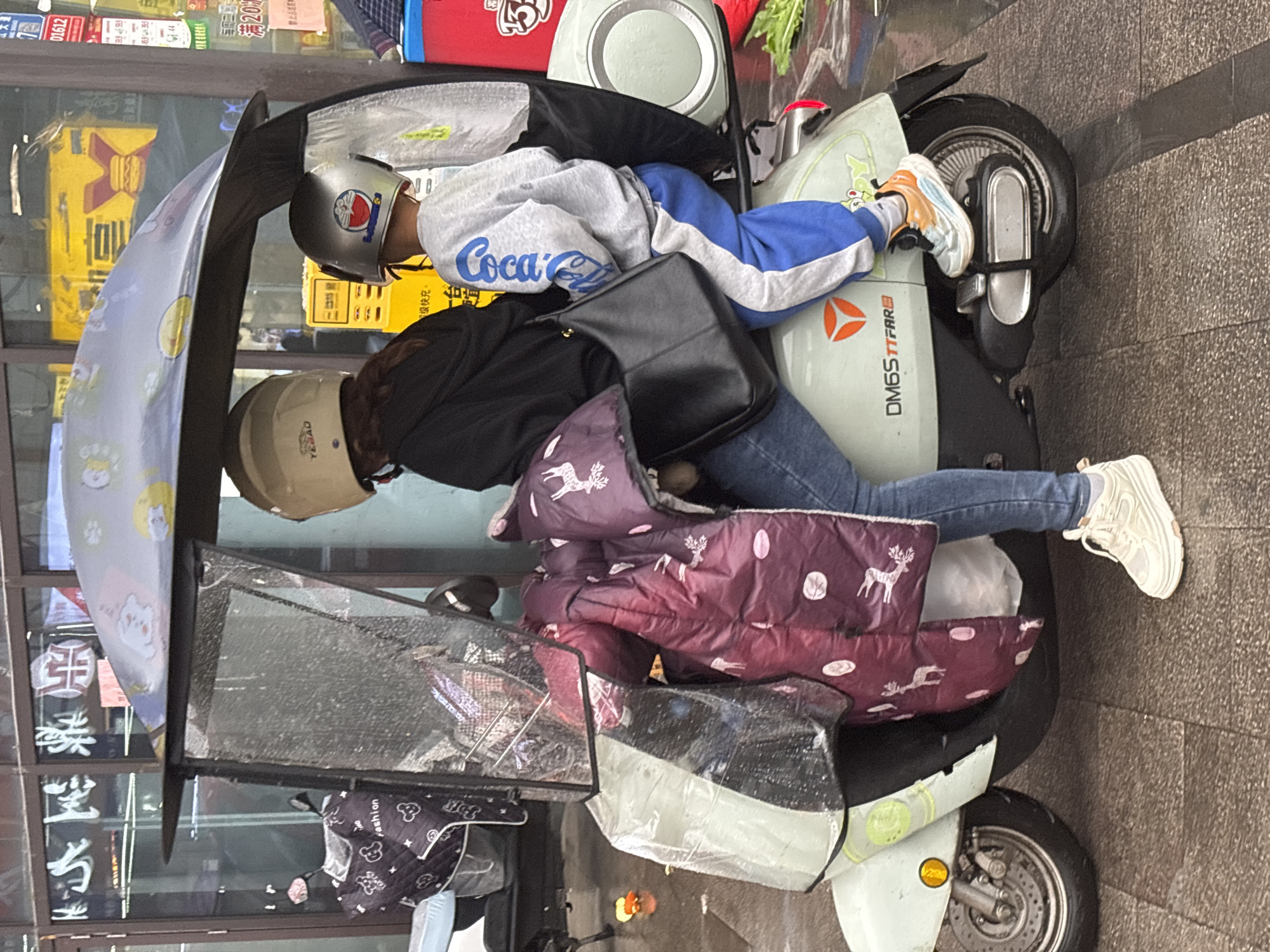 The image shows two people on an electric scooter in Jiangjin, China. The rider, wearing a tan helmet, black jacket, blue jeans, and white sneakers, is seated on the scooter. In front of them is a child wearing a silver helmet with a Doraemon sticker, a grey Coca-Cola sweatshirt with blue stripes, blue pants, and light orange sneakers. The scooter has an attached windscreen with cat cartoon images, a burgundy seat cover with deer patterns, and a black bag hanging on the side. The scooter's license plate reads "DM6S TTFARE". Background details include a storefront with signs and posters in Chinese characters, including what looks to be a hamburger ad. Text on the signs also reads "Coca" and "Fashion". The pavement indicates an urban sidewalk.