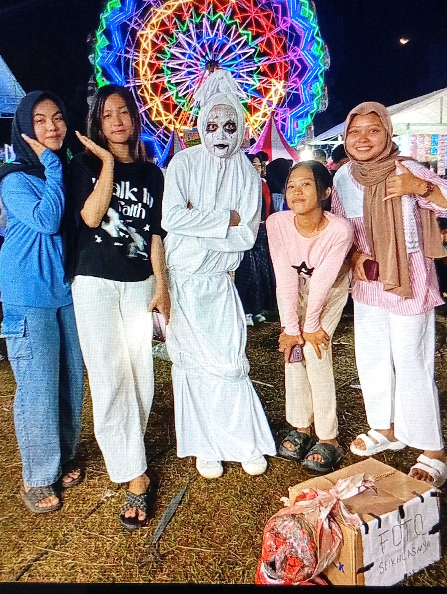 A group of five young women and one individual in a ghost costume pose for a photo at night in front of a Ferris wheel with colorful lights. The Ferris wheel is the main attraction, dominating the background with its vibrant illumination. The group is gathered on a grassy area, possibly a fairground or public space, with a cardboard box in the foreground that has "FOTO" and "SEIKHLASNYA" written on it. One of the women is wearing a hijab and a blue shirt with cargo pants. Another woman is wearing a black t-shirt with white text and white pants. The person in the ghost costume is fully covered in white fabric, with white face paint and black accents around the eyes and mouth. The third woman from the left is wearing a light pink long-sleeved shirt and light-colored pants. The woman on the far right is wearing a hijab and a pink and white striped shirt with white pants. Her right hand is giving a thumbs-up gesture. The environment suggests a festive atmosphere, likely a night market or carnival event. The presence of the Ferris wheel and the illuminated stalls in the background strongly indicate such a setting. The scene appears to be located in Lhokseumawe, Indonesia, given the text on the box which translates to "Photo - As you wish," a common phrase at such events.