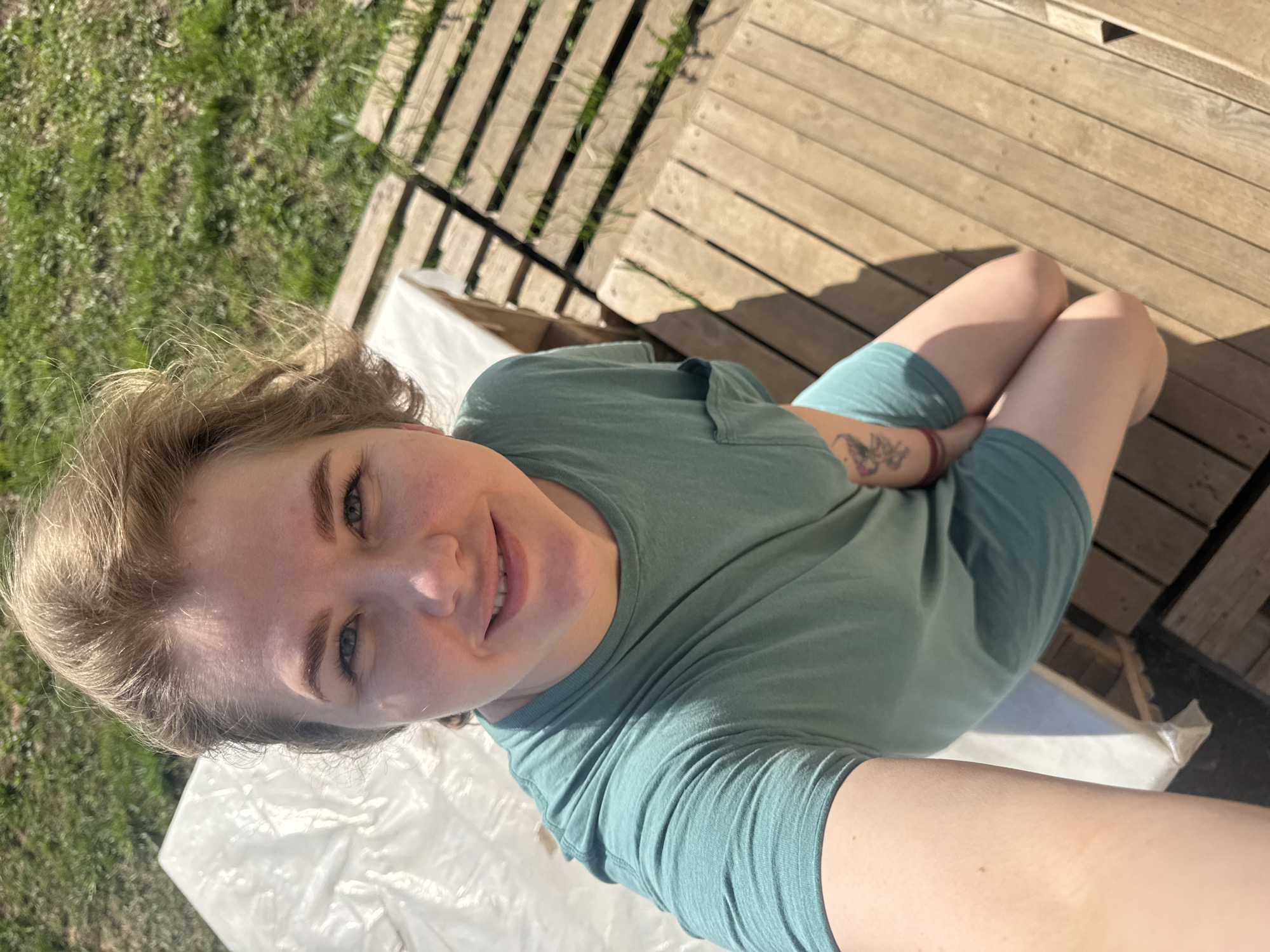 A woman is taking a selfie outdoors in a sunny location. She has fair skin, blonde hair, and is wearing a teal t-shirt and shorts. A tattoo is visible on her left arm. She is kneeling on a white sheet that appears to be covering something. Behind her is a wooden deck or platform. To the left, there is grass. The city of Ферма, Russia is not visibly identifiable in the image.