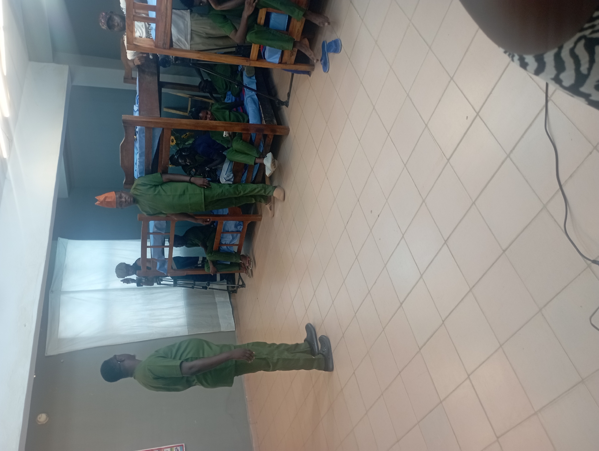 In an indoor room with tiled flooring and white overhead lighting, a group of individuals are present. The room appears to be a dormitory or living quarters, with several bunk beds lining the walls.
On the left side of the image, a man in a green uniform and dark shoes stands facing away from the camera, addressing a group. To his right, near a window covered by white curtains, another man in a similar green uniform stands next to a tripod with a camera.
In the center and right of the image, several people are seated or lounging on the bottom bunks of the wooden bunk beds. They are also wearing green uniforms, with some individuals wearing caps or hats. The beds have blue mattresses and bedding. One person on a lower bunk is looking at their phone. Another individual on the far right appears to be wearing a white shirt and tan pants, leaning against a bunk bed.
The environment suggests a communal living space, possibly for a school, training facility, or similar institution. The overall lighting is bright and even.