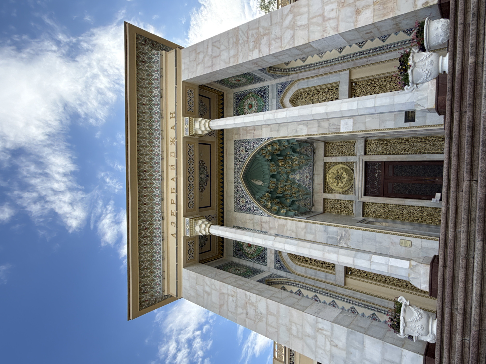 The image shows the Azerbaijan Pavilion at VDNKh (Exhibition of Achievements of National Economy) in Moscow, Russia. The pavilion is a grand, ornate building with marble cladding, intricate mosaics, and gilded architectural elements. The facade features large columns supporting an elaborately decorated entablature with inscriptions in Cyrillic, reading "АЗЕРБАЙДЖАН" (Azerbaijan). Above the entrance, a deep arched recess is adorned with muqarnas (stalactite-like vaulting) and gilded carvings. The entrance itself is a dark wooden door set within a richly decorated portal. On either side of the entrance, tall, narrow windows with golden lattice work are visible. Two white decorative planters with colorful flowers are placed on the marble steps leading up to the pavilion. The sky above is blue with scattered white clouds. The perspective is from a low angle, looking up at the building. There are no people visible in the image.