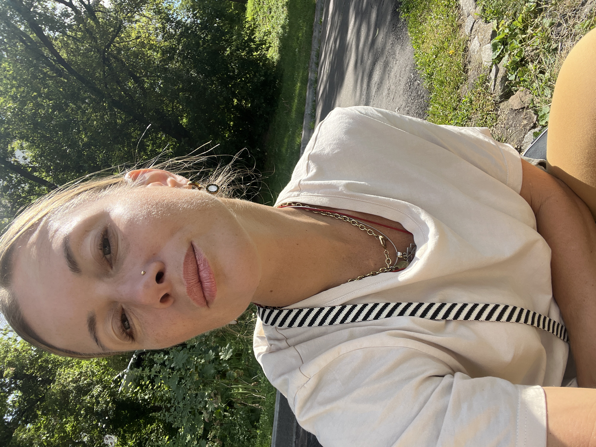 The media file is a close-up, portrait-style selfie taken outdoors during the daytime, likely late afternoon or early evening, given the warm, directional sunlight casting shadows. The subject is a woman with light blonde hair, wearing a cream-colored t-shirt. She has a delicate gold nose stud and appears to be wearing light-colored lipstick. She is adorned with several necklaces, including a gold chain and a red string with a charm. A distinctive black and white striped ribbon is visible across her t-shirt.
The background is blurred but suggests a natural outdoor setting with lush green trees and foliage. A paved path or road can be seen to the right of the subject, with sunlight dappling through the trees onto it. The overall mood is relaxed and casual, with the subject looking directly at the camera with a neutral expression. The lighting creates a slightly backlit effect, highlighting the subject's features and the texture of her skin. The image is slightly tilted, giving it a dynamic, informal feel. No text is visible in the media file.