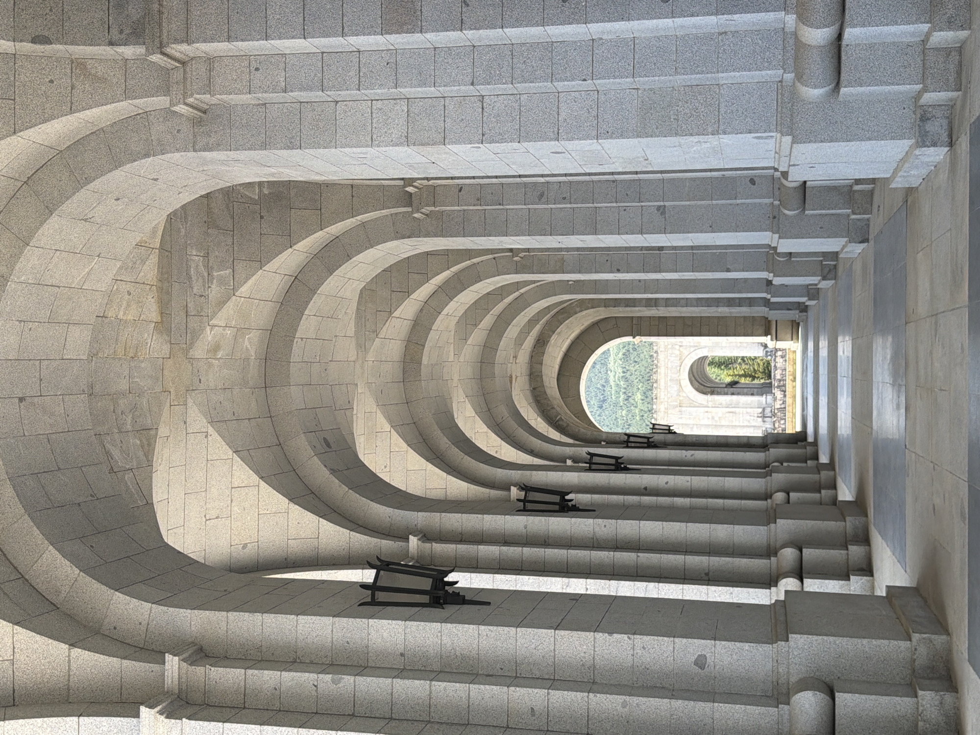 The image shows a perspective view of a long stone colonnade with a series of arches leading into the distance. The architecture is characterized by massive, intricately carved stone pillars and repeating semicircular arches. Black, wrought-iron lanterns are mounted on the pillars at regular intervals. The floor of the colonnade is paved with large, rectangular stones, with some darker square inlays.
In the far distance, through the final archway, a lush green hillside with trees is visible under a clear sky. Another similar stone archway can be seen in the distance, suggesting a continuation of the architectural structure. The light suggests it is daytime, with shadows cast by the arches and pillars, indicating the sun is at an angle. The scene is devoid of any people or animals.
This architectural feature is located within the Royal Site of San Lorenzo de El Escorial, specifically, it appears to be a section of the Cloister of the Four Evangelists or a similar cloistered area within the complex. The Royal Site of San Lorenzo de El Escorial is a historical residence of the King of Spain, located in San Lorenzo de El Escorial, near Madrid, Spain.