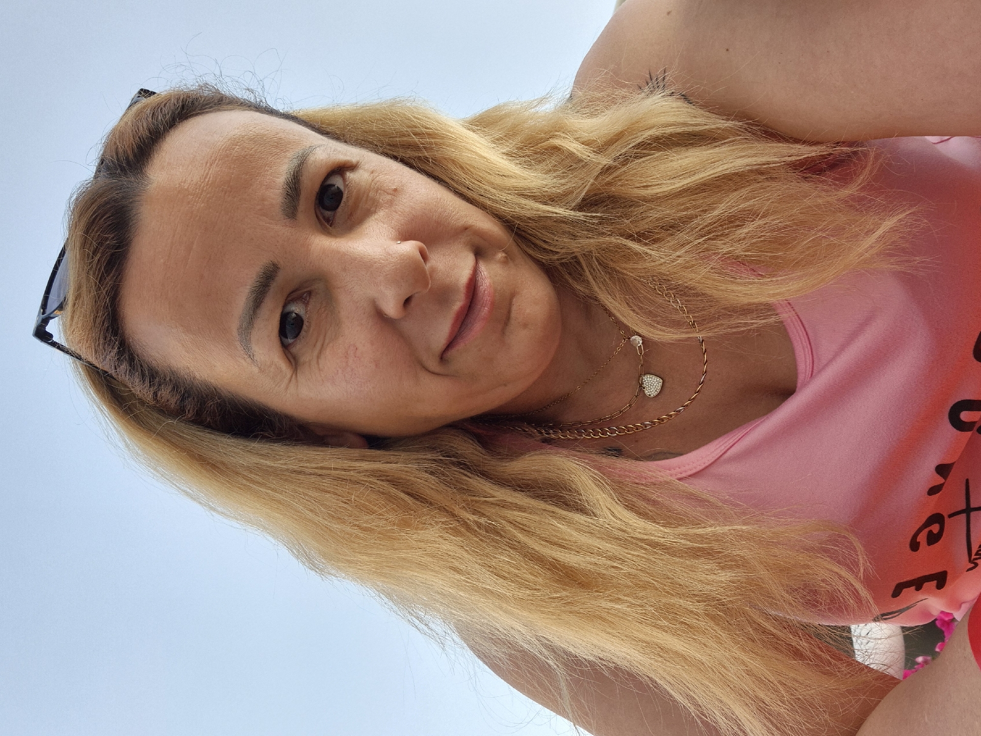 A close-up, low-angle, selfie-style photograph shows a woman with blonde hair, wearing a pink tank top, and gold necklaces, one with a heart-shaped pendant. She has sunglasses perched on her head, and her left arm is visible. The background is a clear, light blue sky. The image was taken outdoors, likely during the daytime, with no identifiable landmarks or city details.