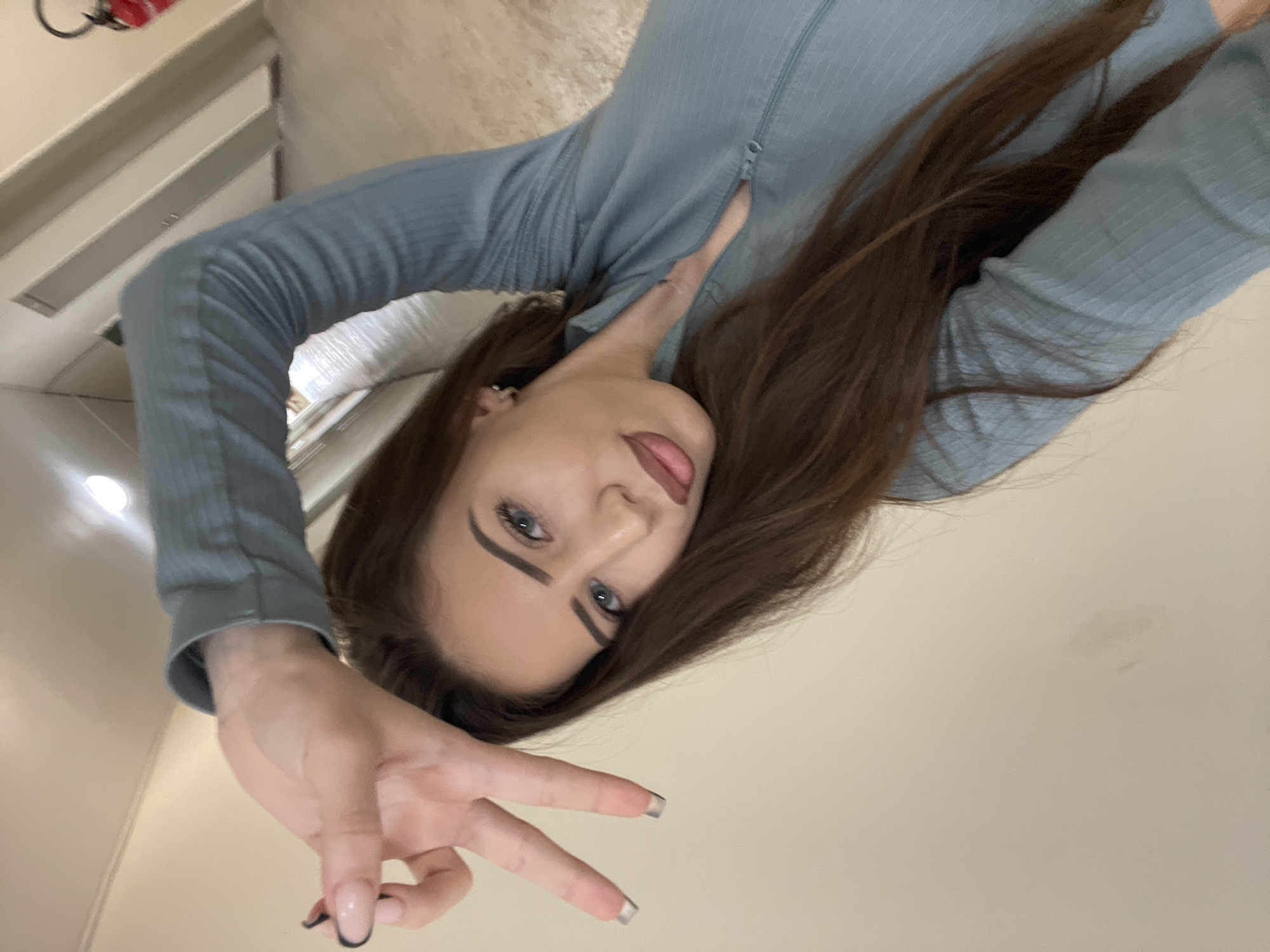 A woman with long brown hair is making a peace sign with her hand. She is wearing a long-sleeved light blue top. The background appears to be a room in a building, with what seems to be a shelf or cabinet visible in the top left corner. The image is taken from a slightly angled, close-up perspective. There is no indication in the image that it was taken in the city of Veliko Tarnovo, Bulgaria.