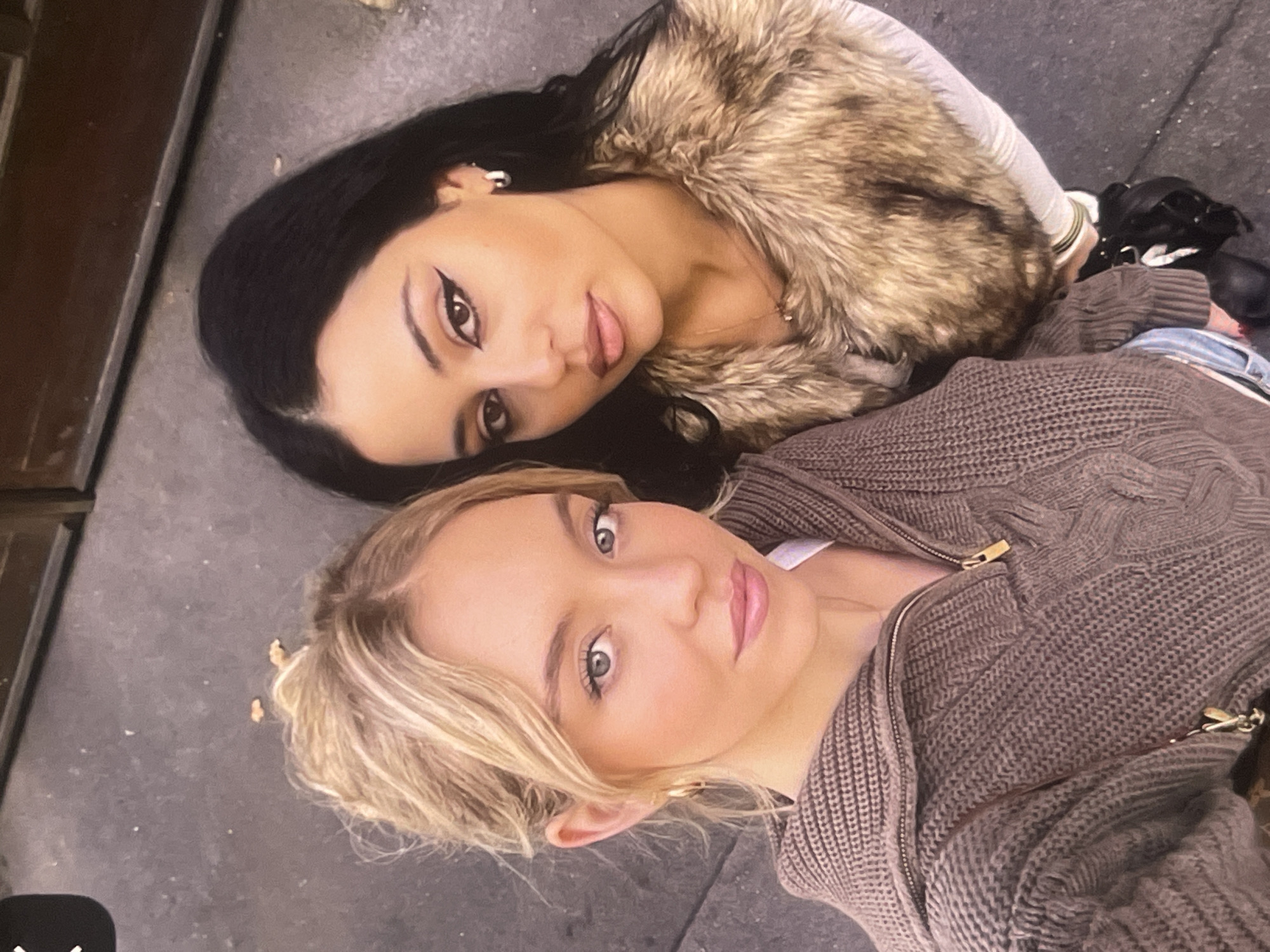 Two young women are captured in a close-up, top-down shot, seemingly lying on a dark, flat surface, possibly pavement or a similar outdoor material. The image has a slight tilt, making them appear to be resting their heads on the ground while looking up at the camera.
The woman at the top of the frame has long, dark hair and striking brown eyes. She wears a winged eyeliner, subtle eye shadow, and light lip gloss. She is dressed in a light-colored top covered by a substantial faux fur vest or jacket with varying shades of brown and beige. She also wears earrings and a delicate necklace.
The woman at the bottom of the frame has blonde hair, light-colored eyes, and natural-looking makeup, including lip gloss. She is wearing a warm, brown, cable-knit cardigan or jacket with a visible zipper and golden hardware. A lighter-colored top is visible underneath her cardigan.
Both women are looking directly at the camera with neutral to slightly smiling expressions, suggesting they are posing for a casual selfie or portrait. Their proximity and relaxed poses indicate a friendly interaction. The dark, textured surface they are on and a dark vertical element on the left side of the frame provide the backdrop. Their clothing suggests cool weather conditions. No discernible text is visible in the image. The setting for this photograph is Poltava, Ukraine.