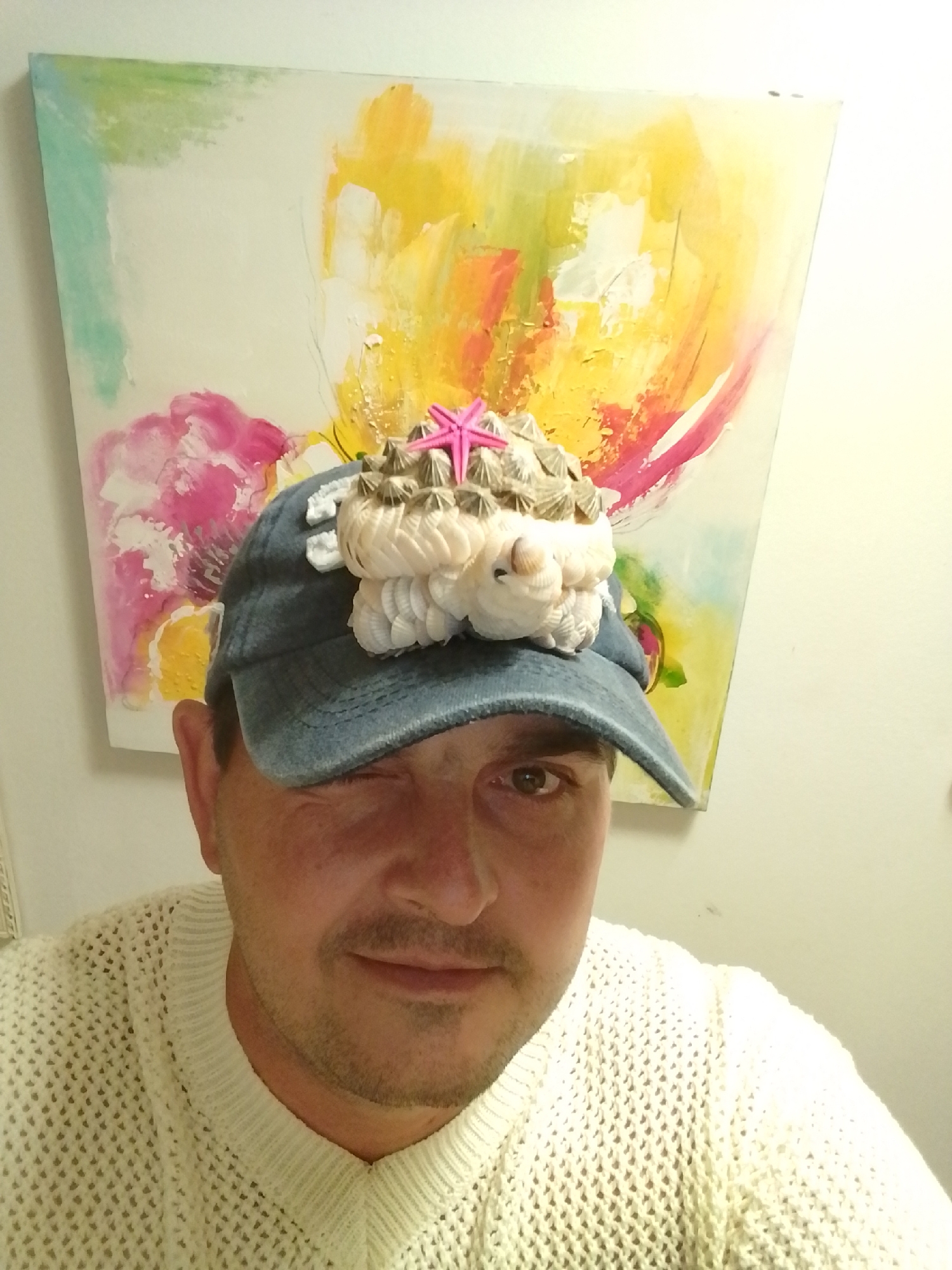 A medium shot shows a person wearing a blue baseball cap decorated with shells and a pink starfish. The cap is adorned with what appears to be a cluster of barnacles and small white shells. The person is also wearing a cream-colored knitted sweater. They are looking towards the camera with their right eye winking. In the background, there is a large, abstract painting with vibrant colors including yellow, pink, and teal. The overall setting appears to be indoors, possibly in a home or studio. No specific location in the city of Wedel, Germany is identifiable from the image itself.